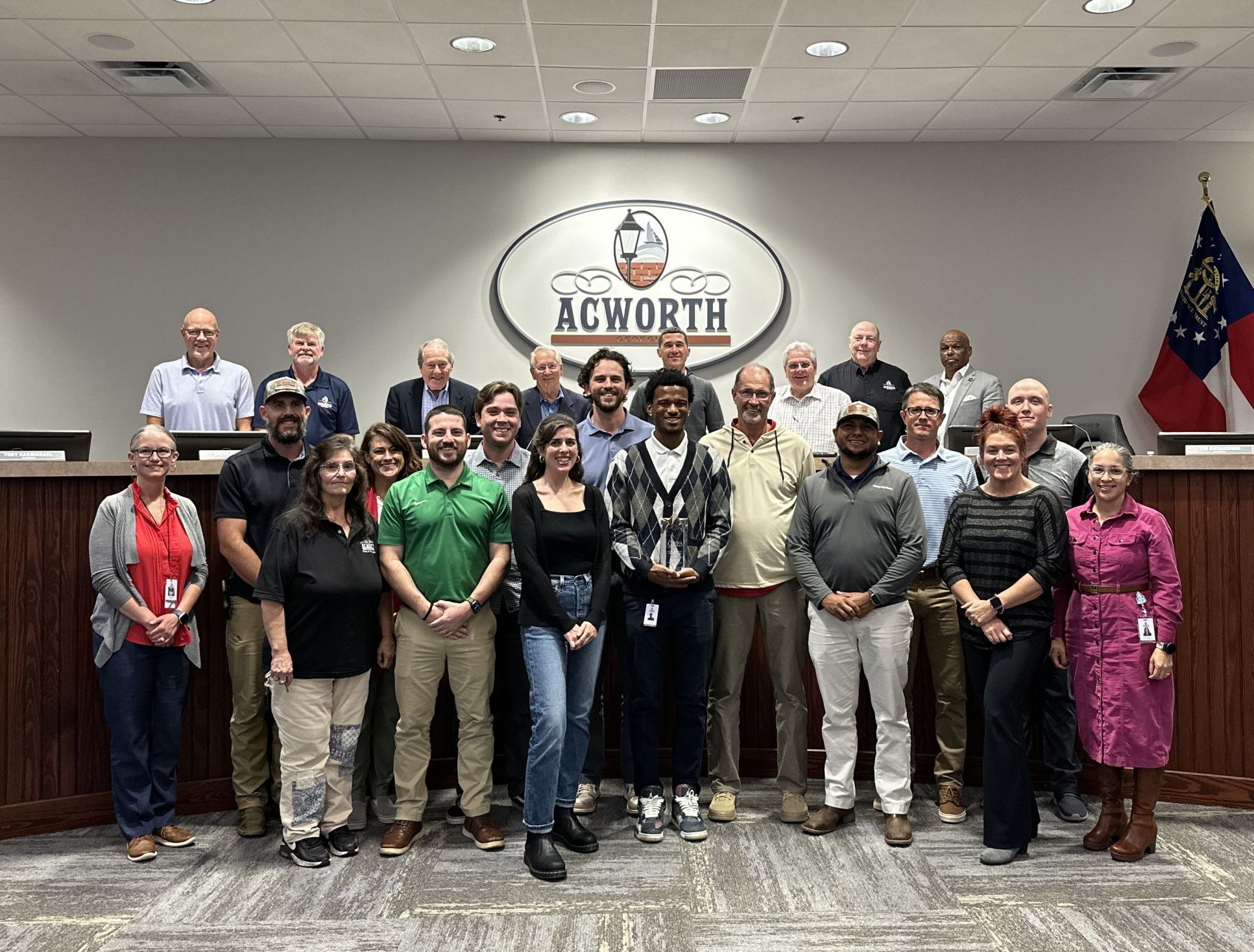 A group of people pose for a photo in a government chamber with an Acworth sign and two flags visible in the background.