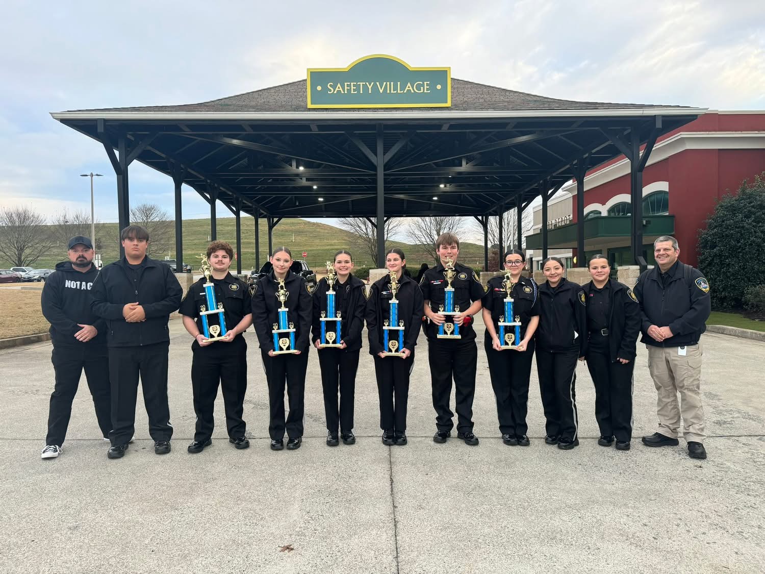 A group of people in uniform stand outdoors under a pavilion labeled "Safety Village," holding trophies and posing for a group photo.