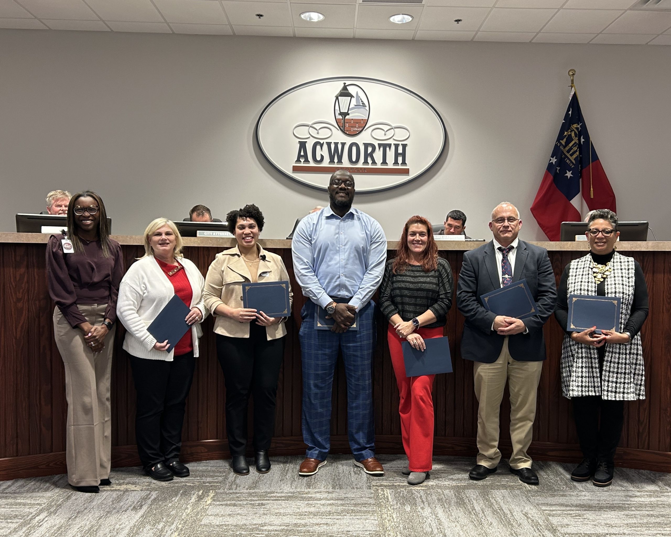 Seven people stand in front of a wooden desk and an Acworth sign, six holding certificates, with the Georgia state flag visible in the background.