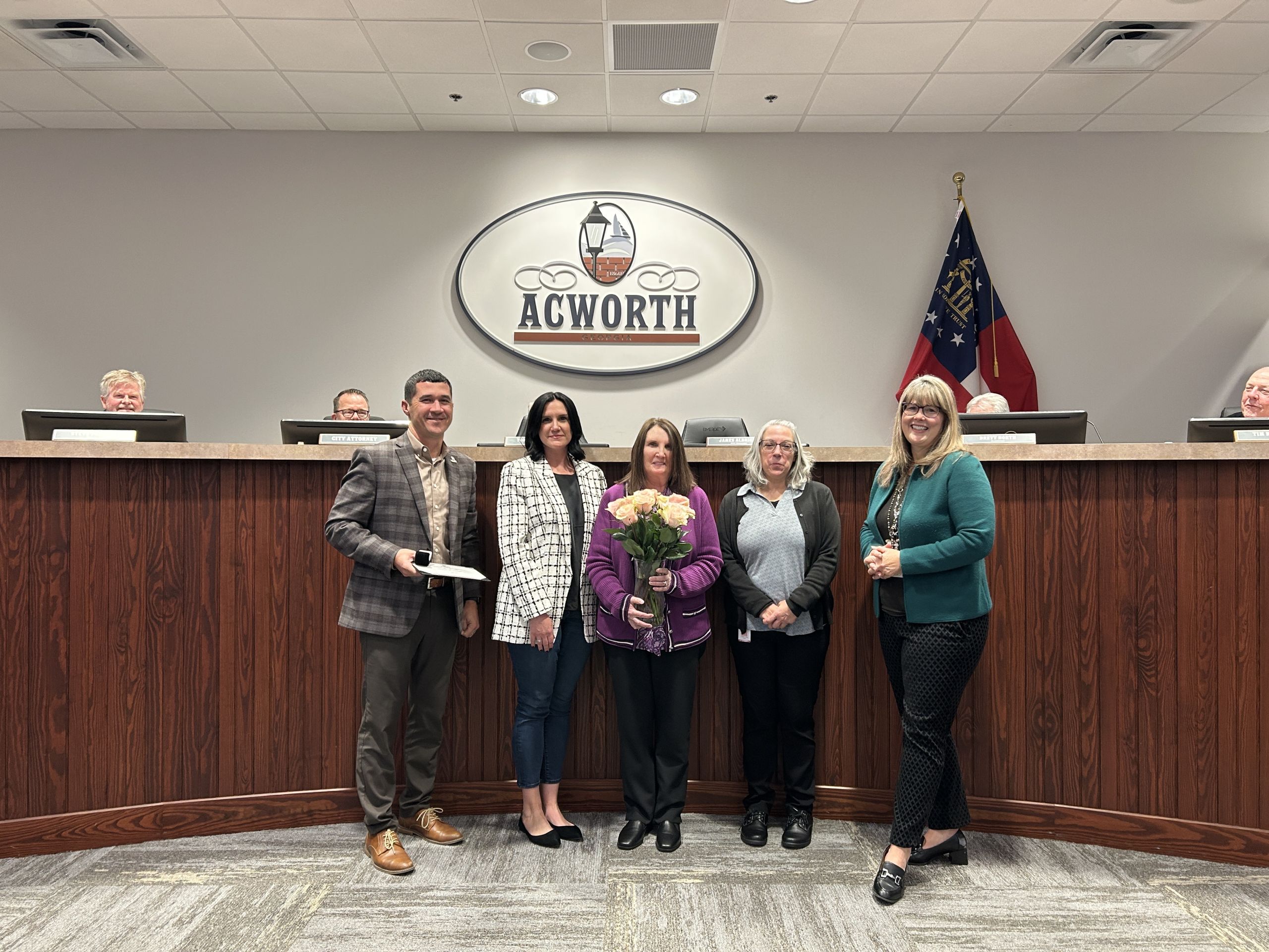 Five people stand and pose for a photo in front of a wooden panel and "Acworth" sign, with some holding flowers; several officials are seated in the background.