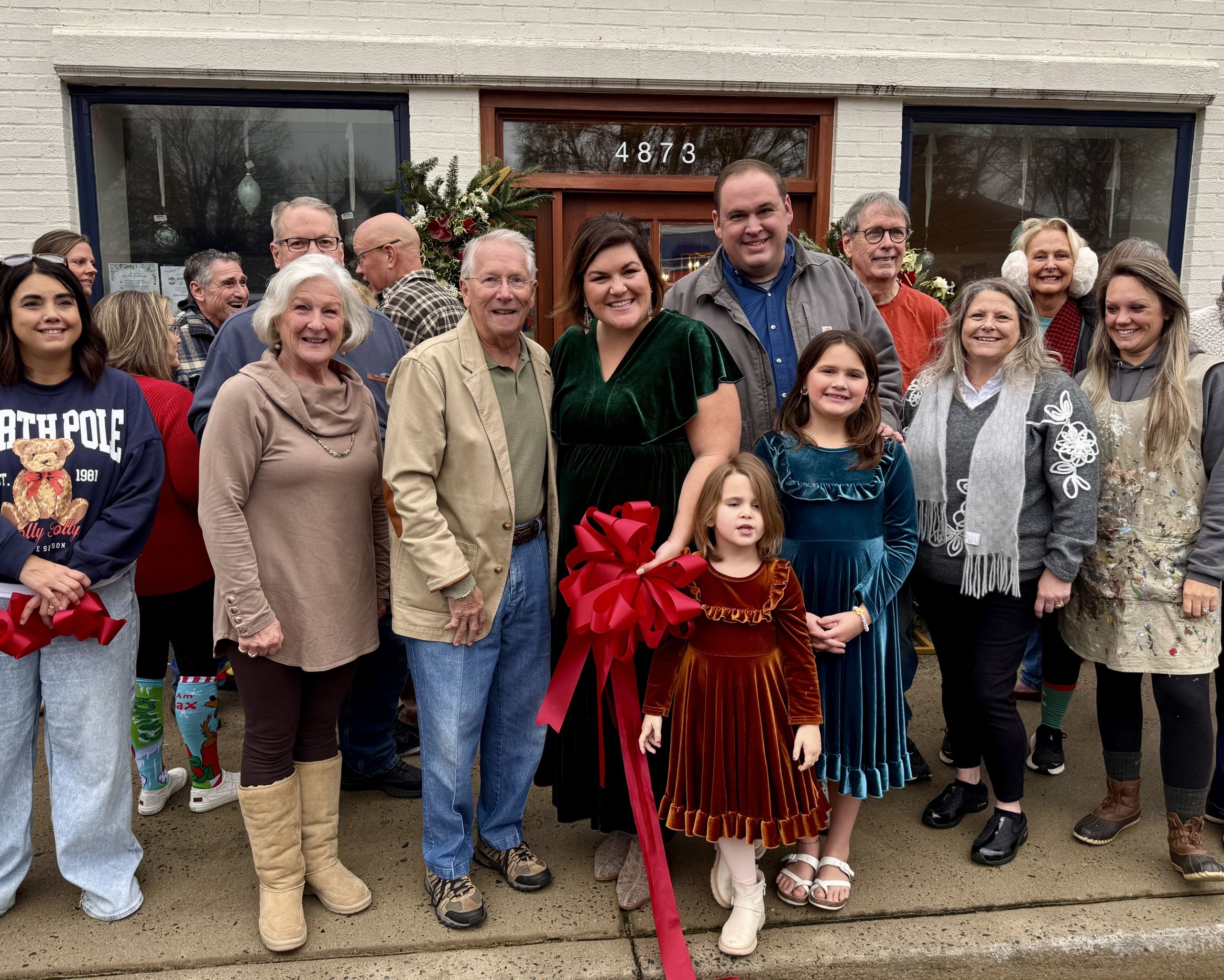 A group of people, including adults and children, stand outside a building with a large red ribbon, suggesting a ribbon-cutting event.