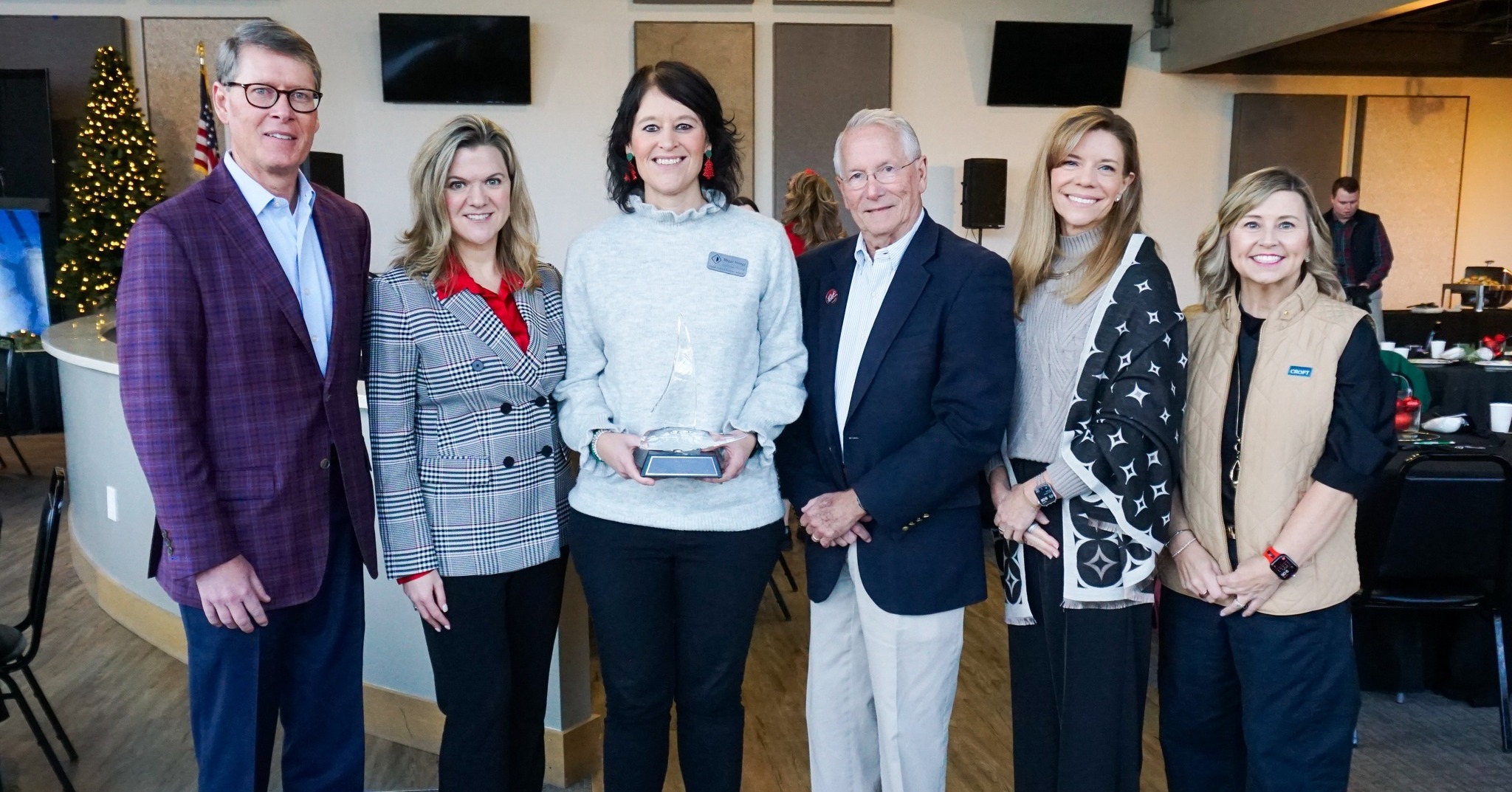Six adults stand indoors in a group, with one woman in the center holding a clear award. There are tables, chairs, and a decorated Christmas tree visible in the background.