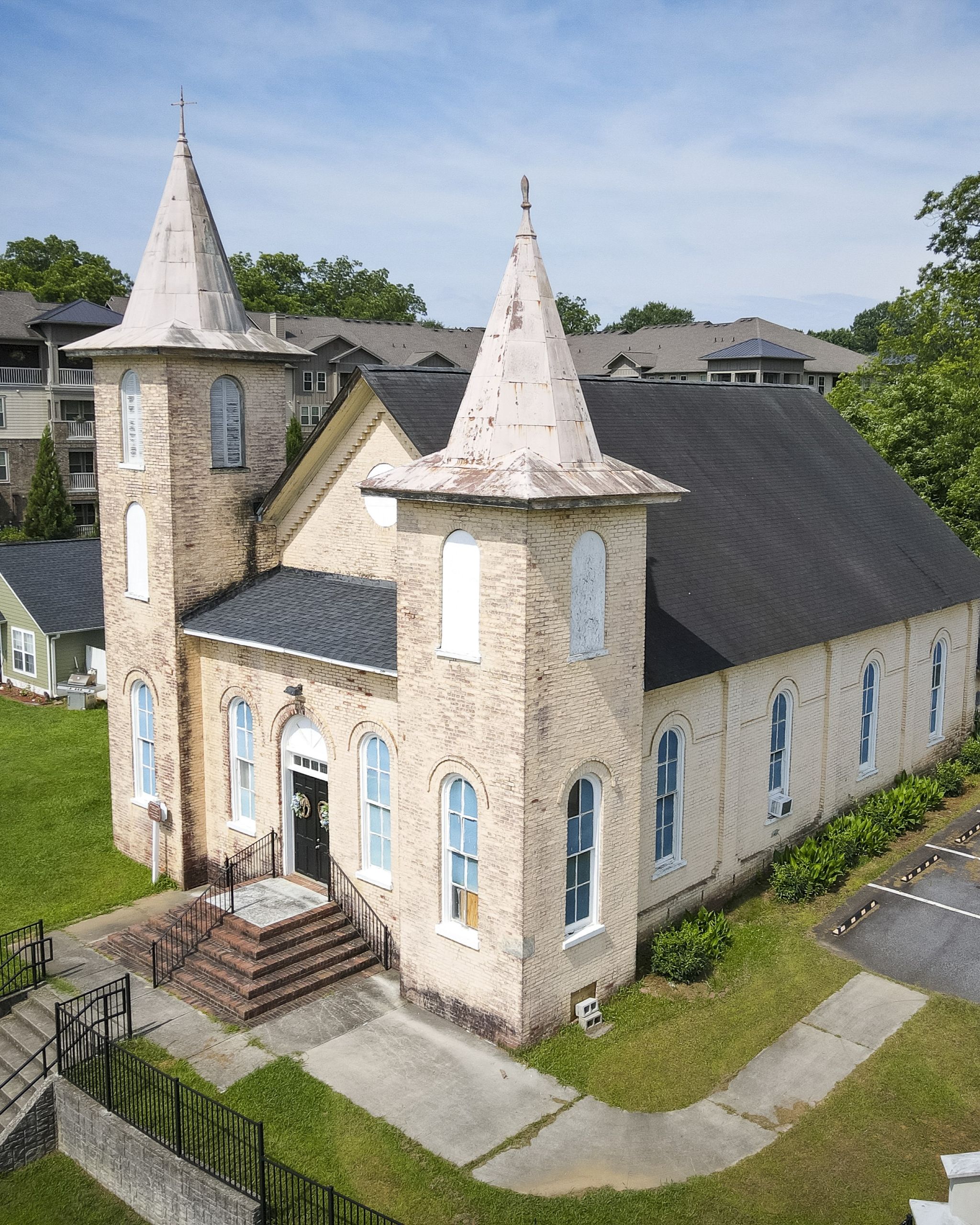 A brick church with twin towers, arched windows, and a dark roof sits on a grassy lot in Acworth, next to a parking area and residential buildings.