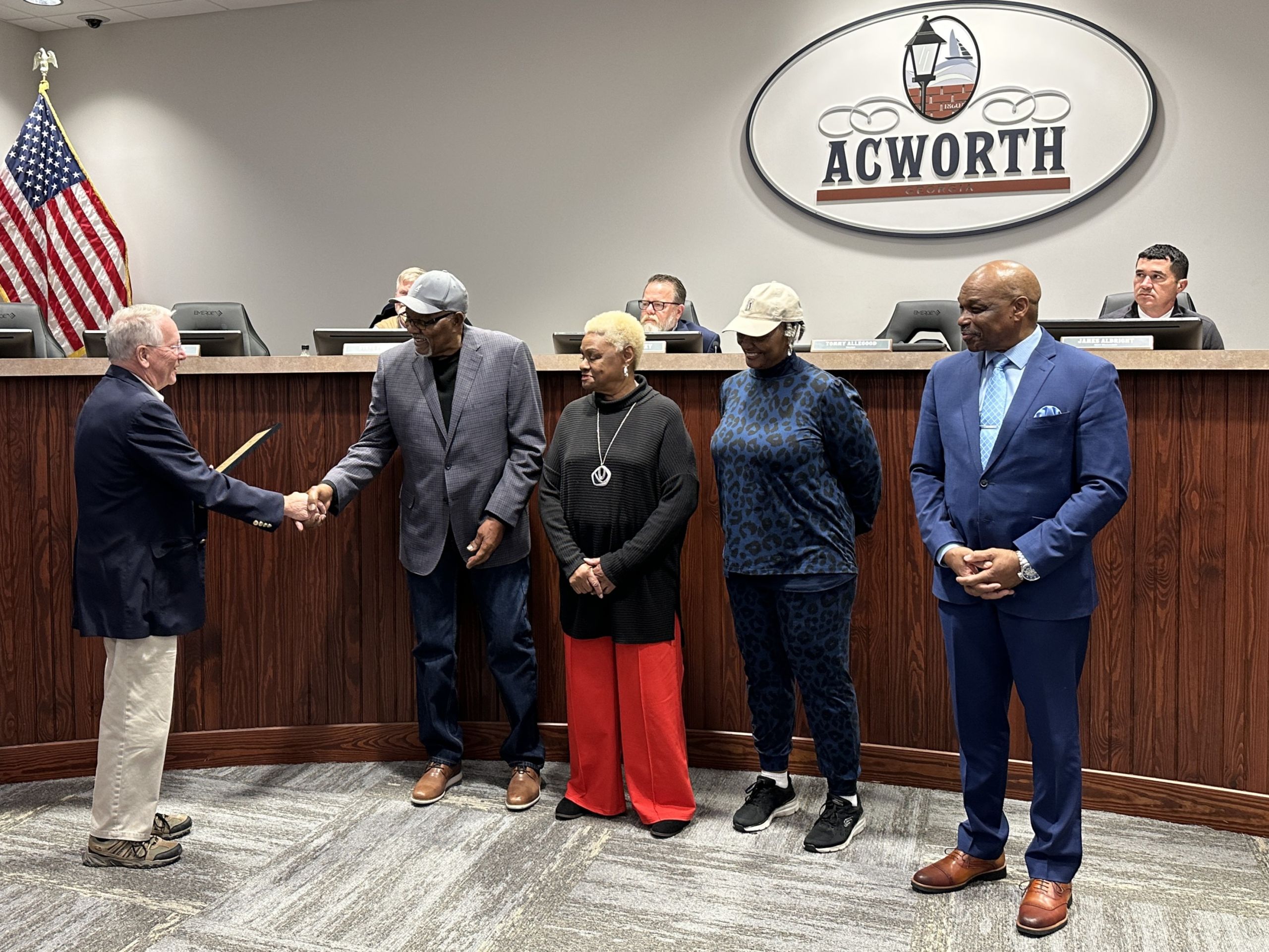 Four people stand at the front of a meeting room as a man presents a proclamation and shakes hands in recognition of Martin Luther King Jr. Day; an American flag and Acworth logo are visible in the background.