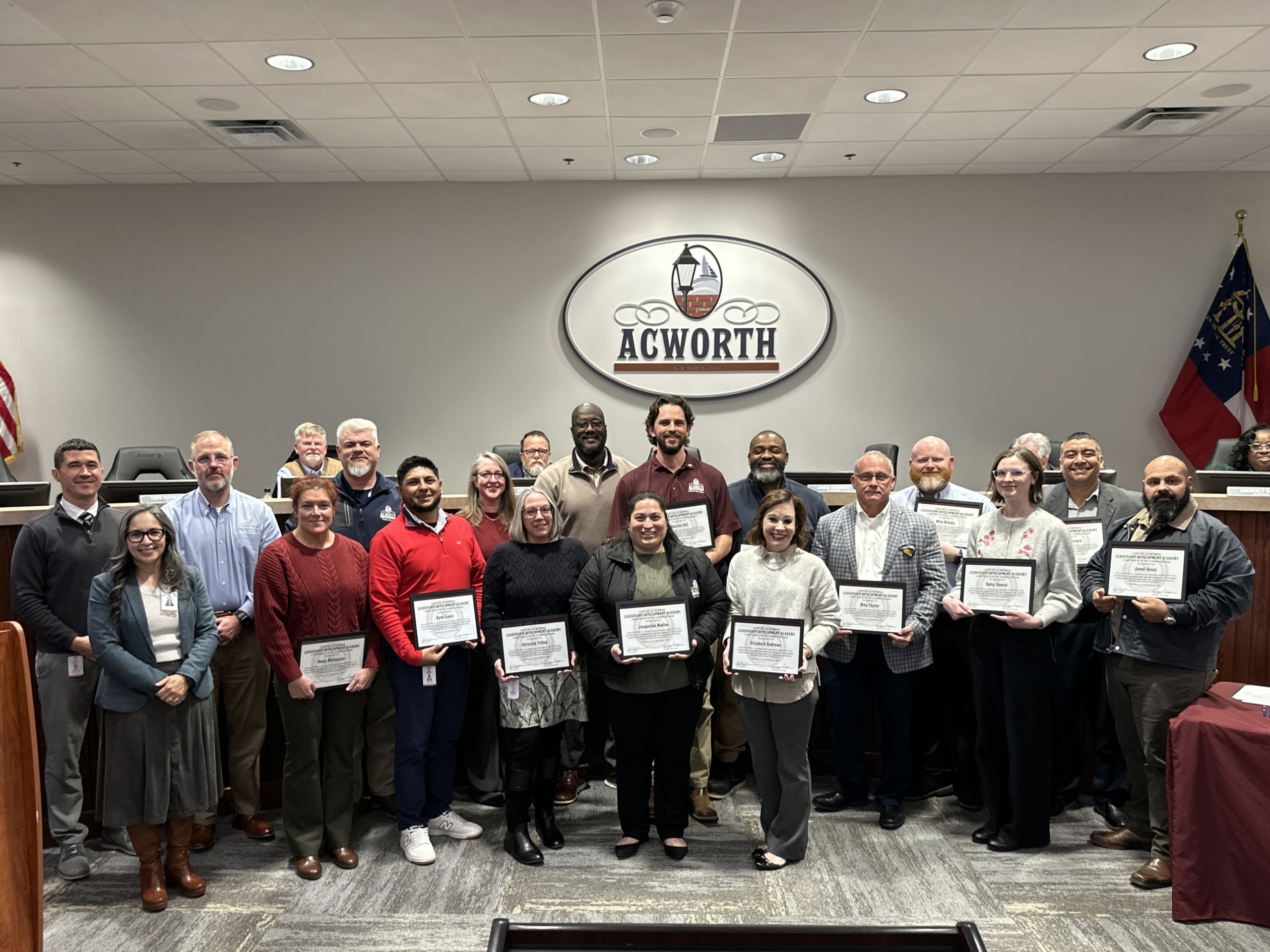 A group of Leadership Development Academy graduates pose indoors holding certificates, standing in front of an "Acworth" sign in a formal setting.