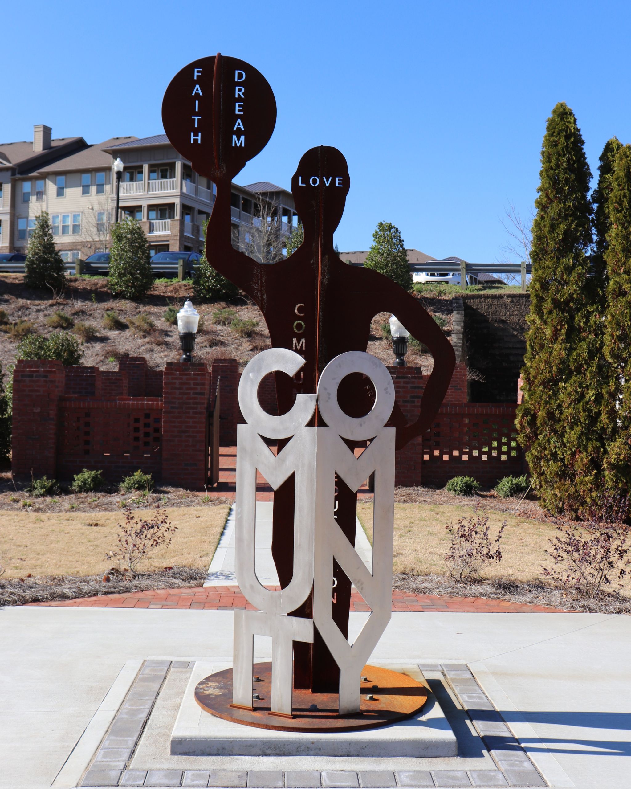 A metal sculpture in Acworth spells out "COMMUNITY," featuring a human figure holding circles labeled "FAITH," "DREAM," and "LOVE." This artwork celebrates unity and Black History amid an outdoor plaza with buildings and trees in the background.
