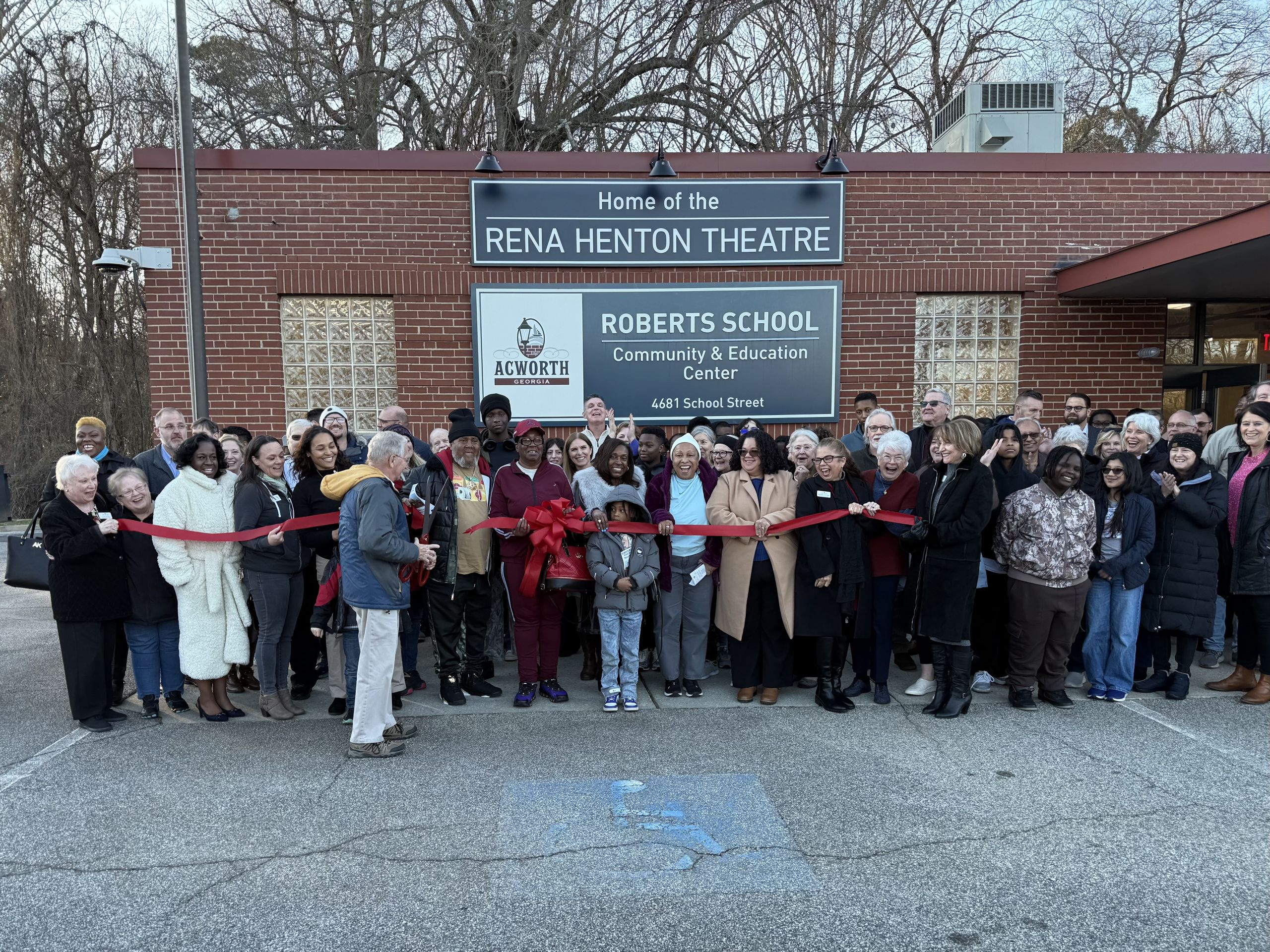 A diverse group of people stands outside the Rena Henton Theatre at the Roberts School, gathered for a ribbon-cutting ceremony in front of the building.