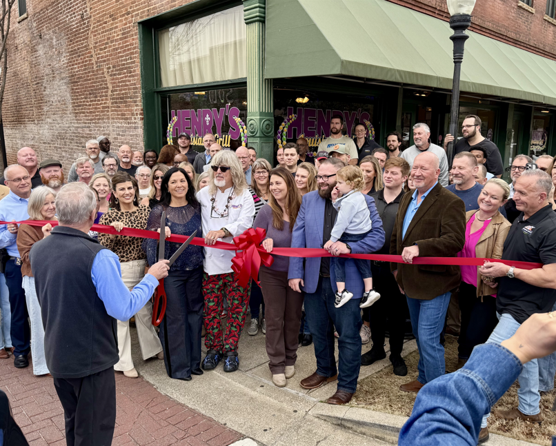 A large group gathers outside Henry's Louisiana Grill as a man cuts a red ribbon for the grand opening Ribbon Cutting ceremony.