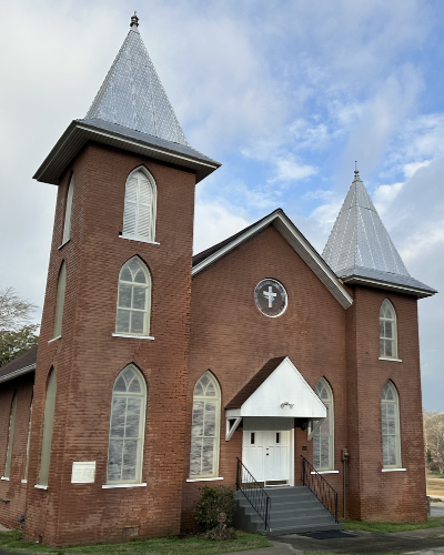 Red brick church building in Acworth with two tall towers, pointed silver roofs, arched windows, front steps, and a cross above the entrance—an iconic site reflecting the area’s Black History on a partly cloudy day.