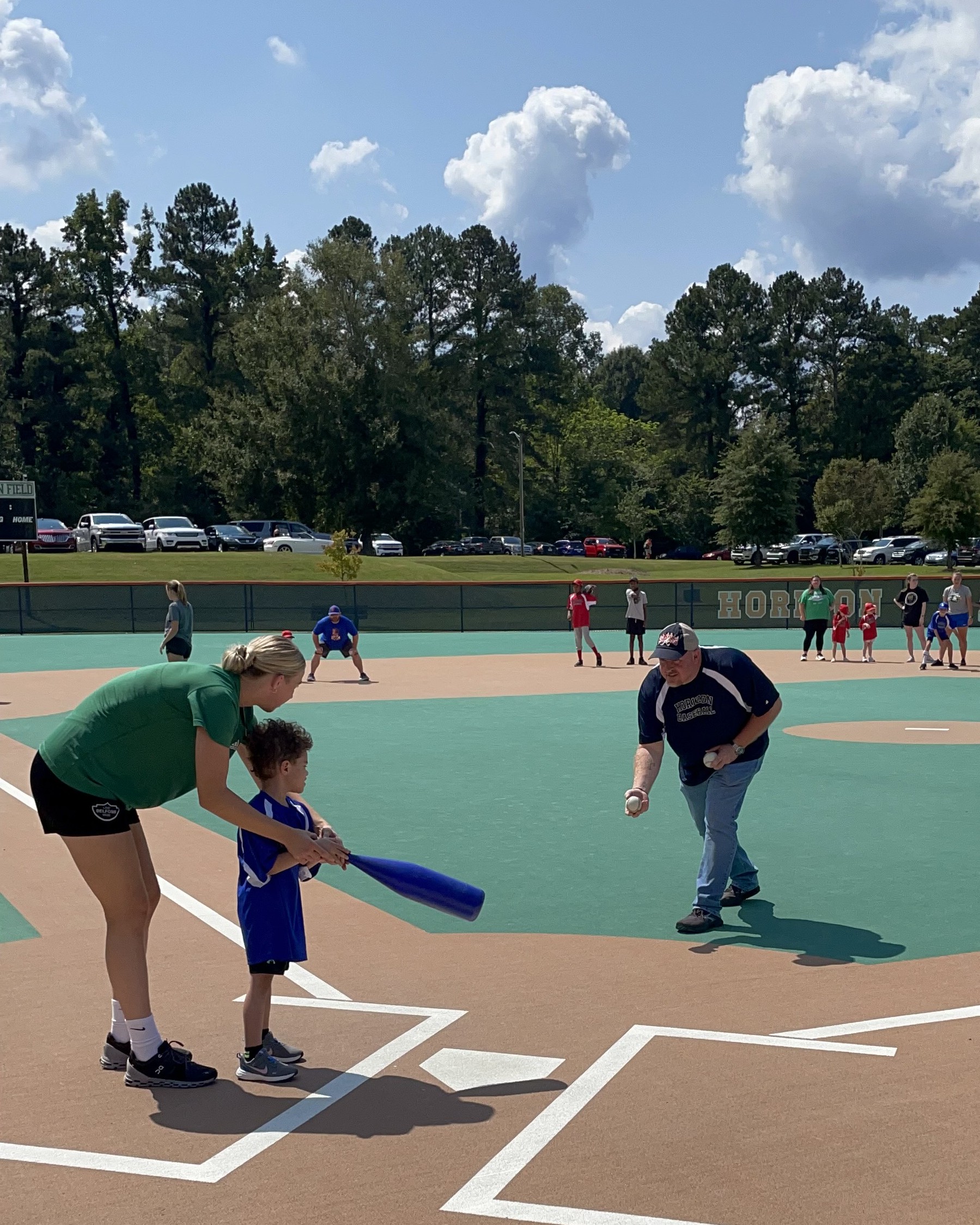 IMG_0167 An adult helps a young child holding a blue bat prepare to hit while another adult pitches during a baseball game on a sunny day, creating a perfect moment for the Horizon Blog.