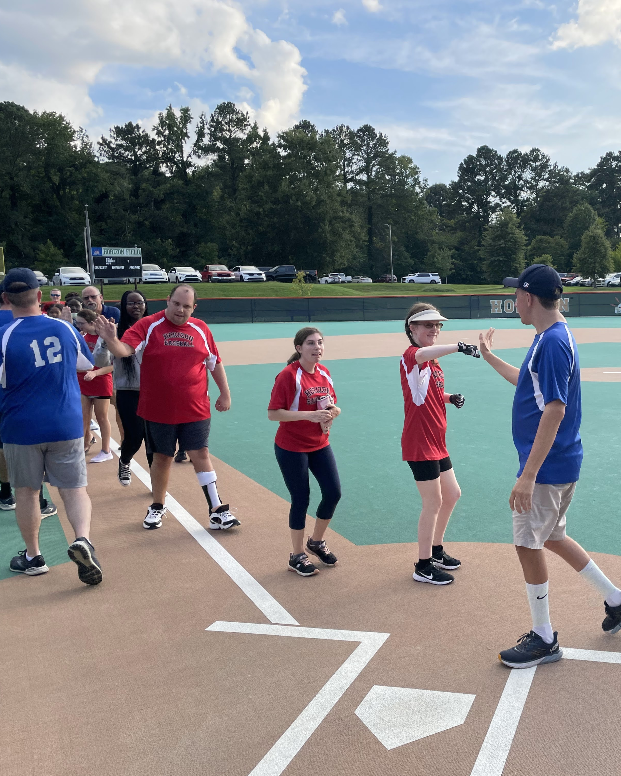 IMG_1708 Players in red and blue shirts line up and shake hands on a baseball field, with trees, parked cars, and a partly cloudy sky in the background—a picture-perfect moment for the Horizon Blog.