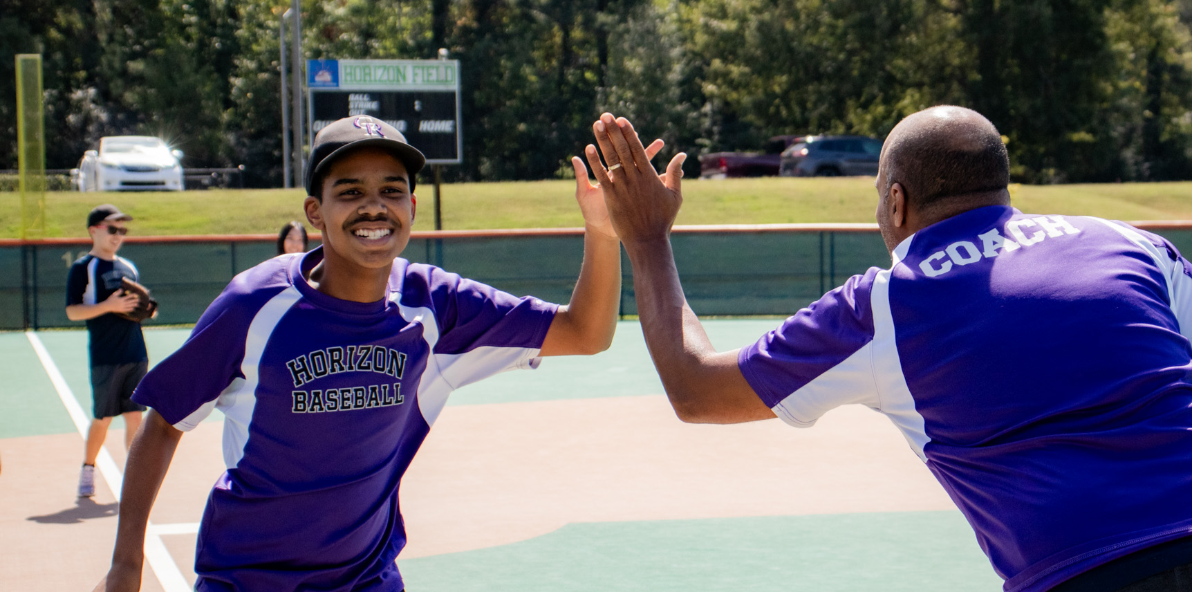 A baseball player and a coach in purple "Horizon Baseball" shirts share a high-five on the field, capturing a winning moment for the Horizon Blog.