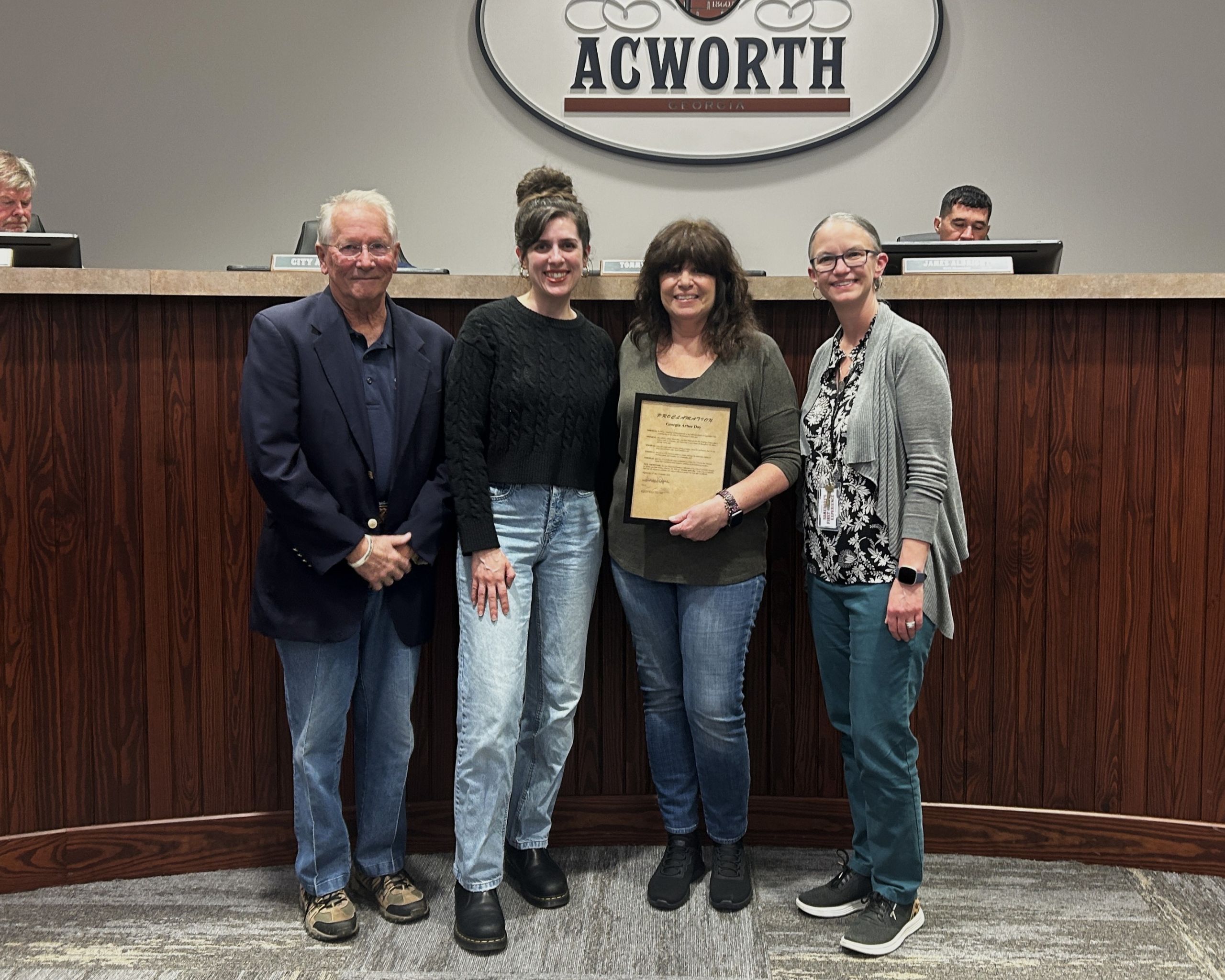 Four people stand indoors, one holding a plaque, in front of a panel beneath a sign that reads "Acworth," celebrating Georgia Arbor Day with a special proclamation.