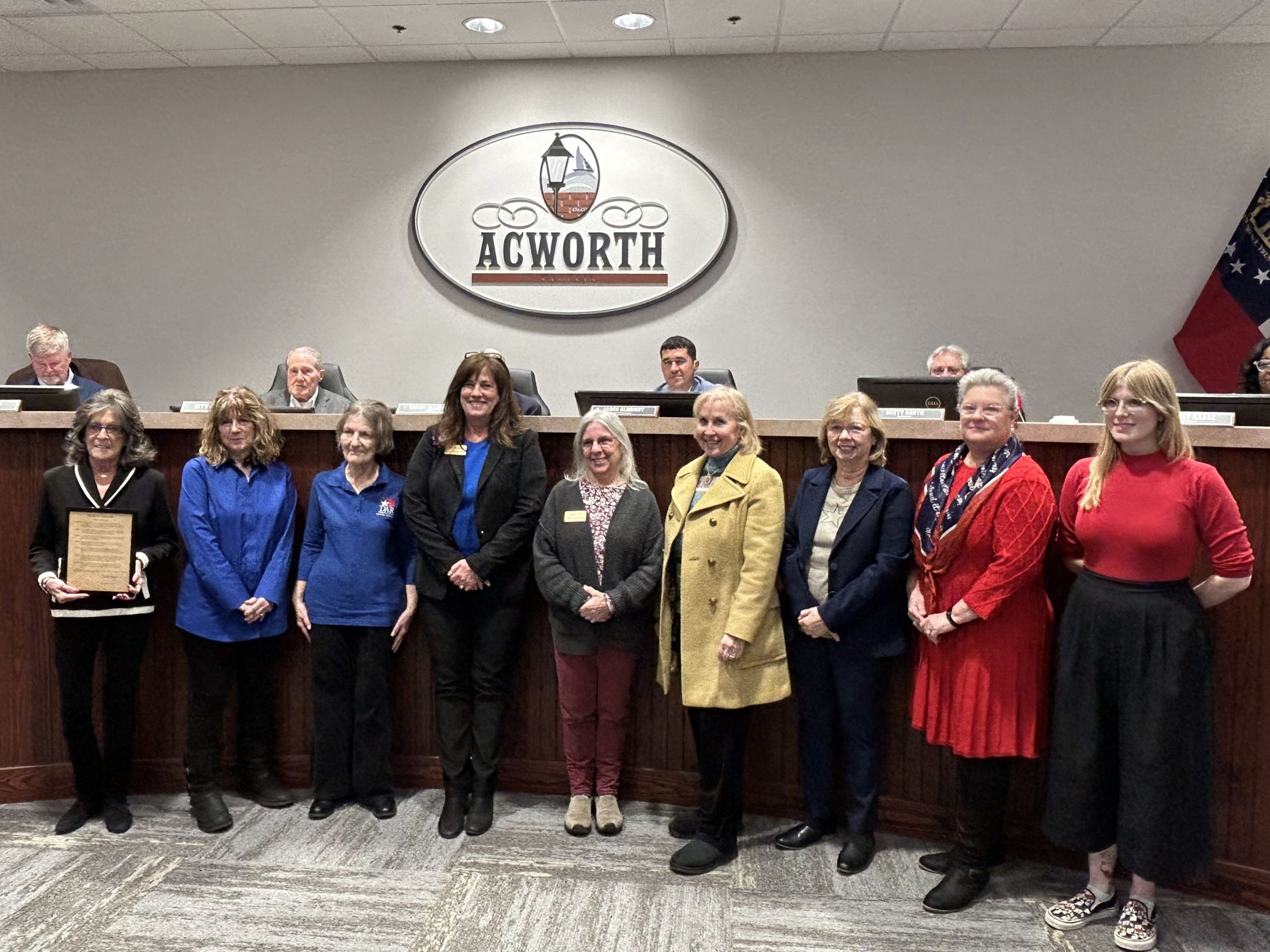 A group of women stands in front of a city council bench with officials seated behind them under an "Acworth" sign; one woman holds a plaque, celebrating a proclamation and recognition on Georgia Day.