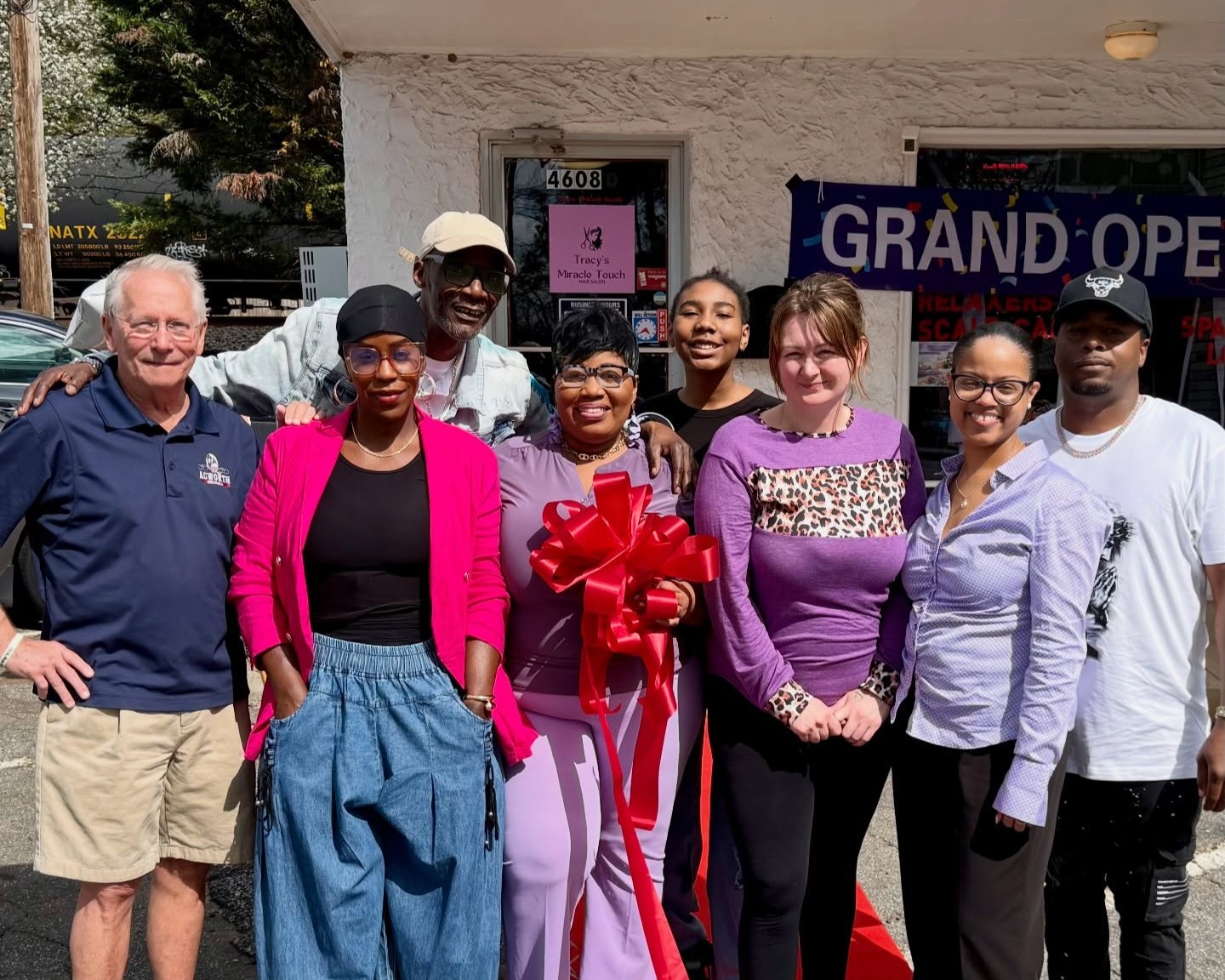 A group of eight people stand together outside Tracy’s Miracle Touch Hair storefront with a "Grand Opening" banner, one person holding a large red ribbon bow in celebration of the ribbon cutting and hair celebration event.