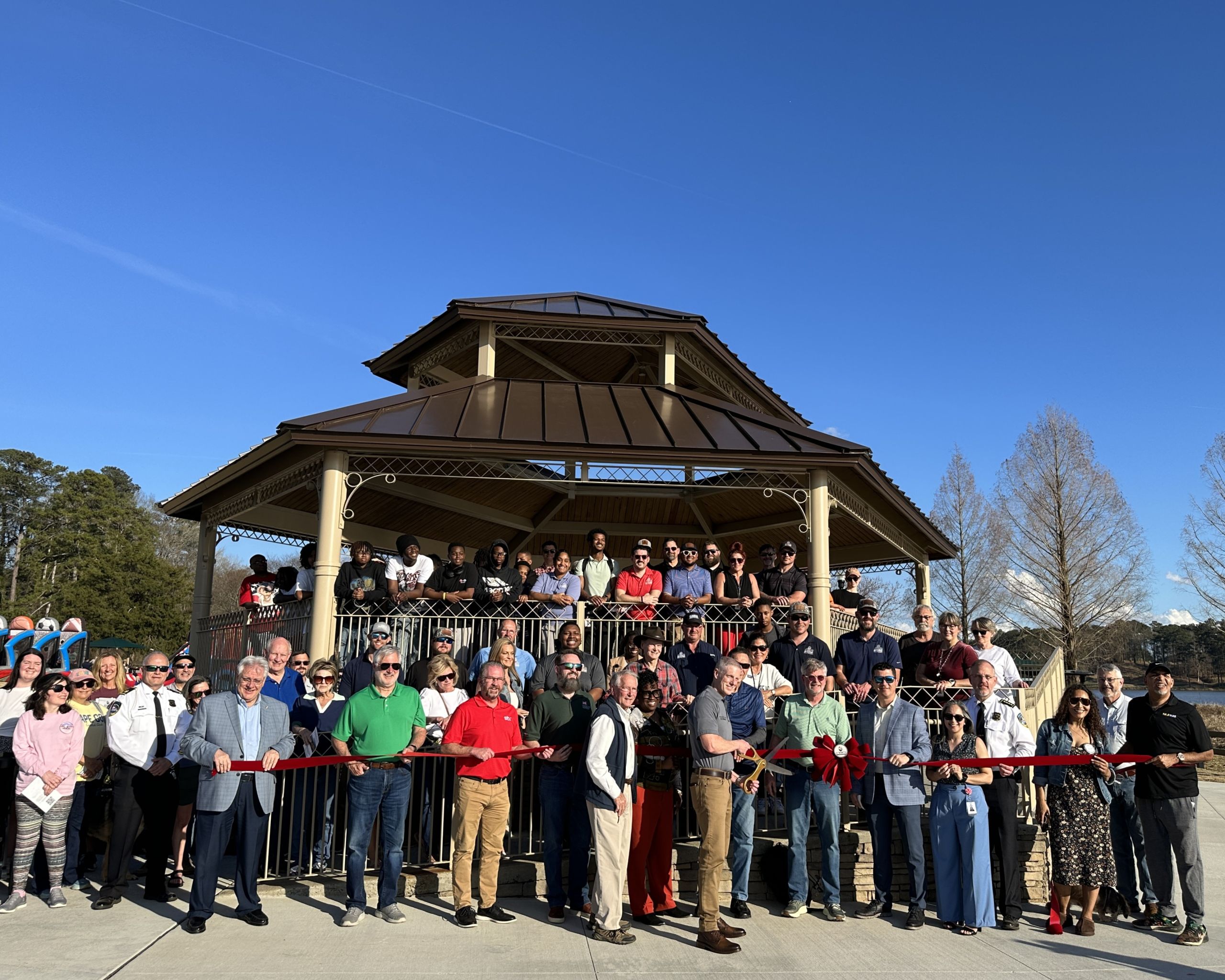 A large group gathers in front of a gazebo at Lake Acworth for a Master Plan ribbon cutting on a sunny day; several individuals hold a large red ribbon across the front.