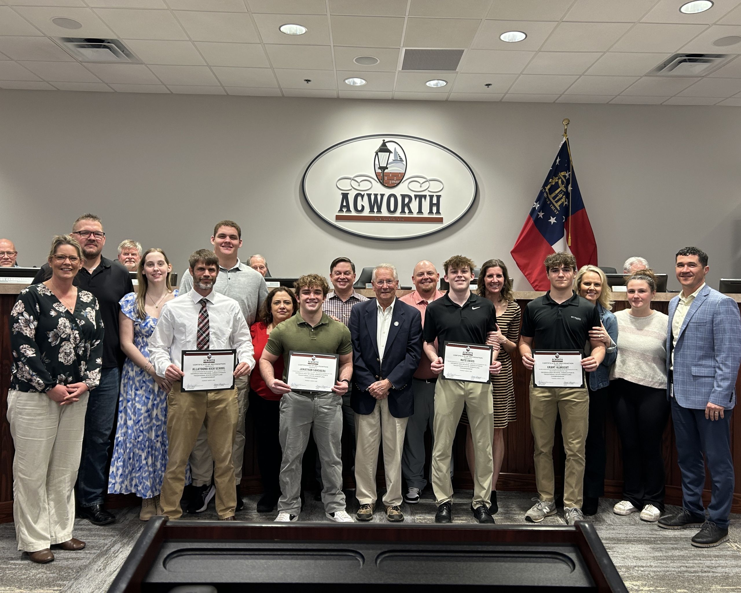 A group of people, including members of the Allatoona High School Wrestling Team, pose indoors at Acworth City Hall for a recognition event, with certificates in hand and the Georgia state flag and Acworth logo in the background.
