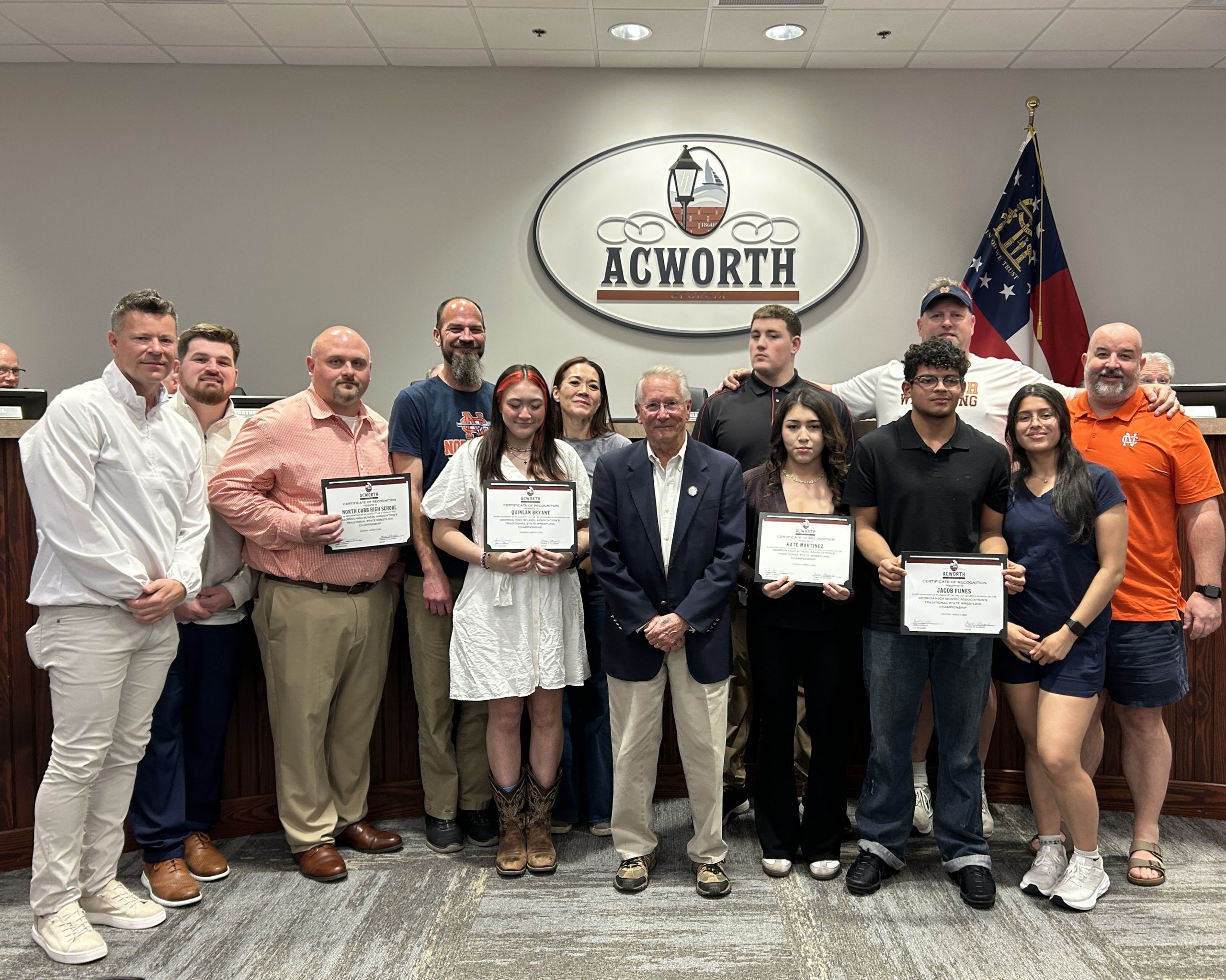 A group of people pose indoors, some holding certificates, in front of an Acworth sign and a Georgia state flag, celebrating achievements in North Cobb Wrestling.