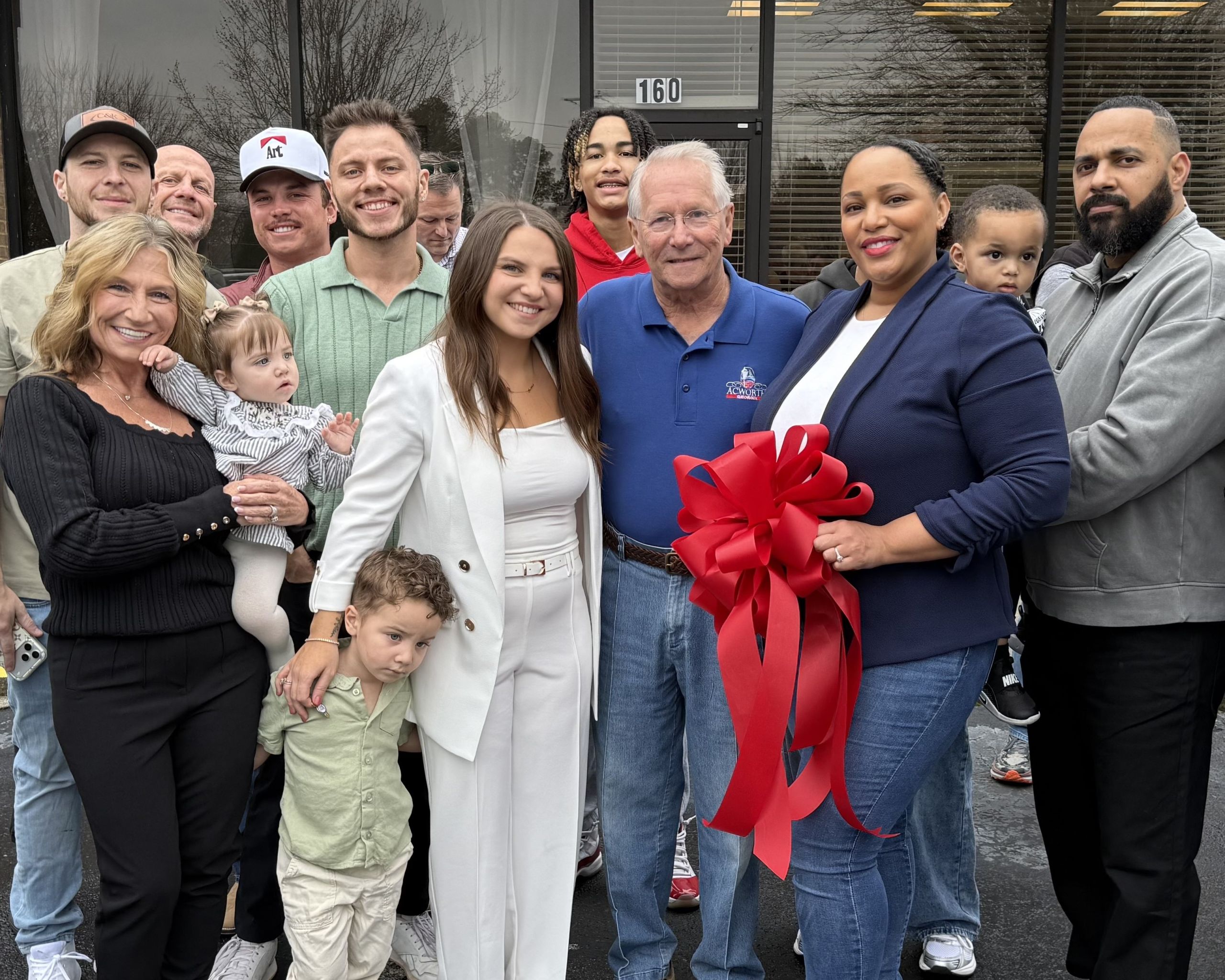 A group of people stands outside The Suites by Jade’s entrance, celebrating a ribbon cutting. One woman holds a large red ribbon bow as adults and children gather together, smiling for a group photo.