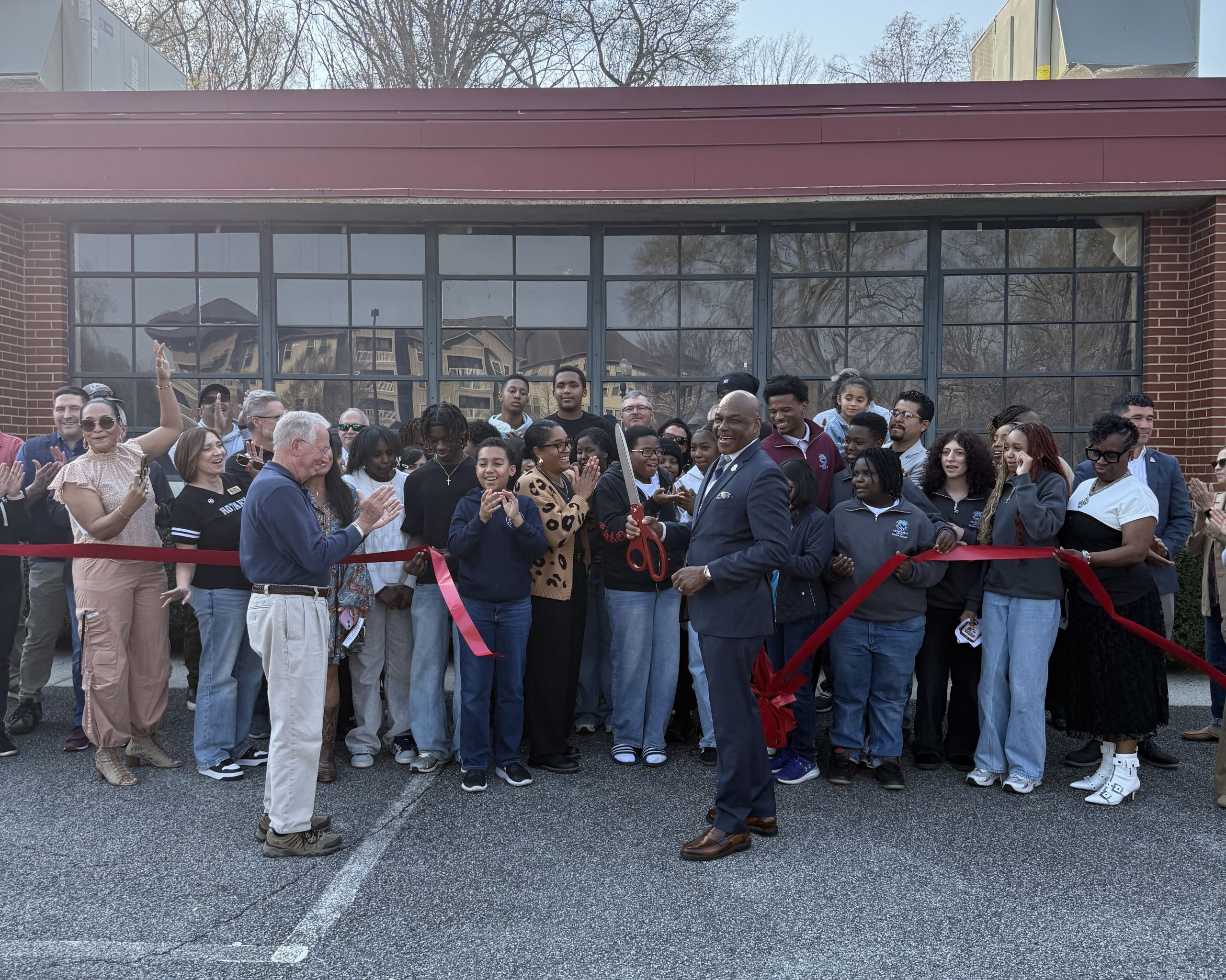 A group of people stand outside a building, gathered for a ribbon-cutting ceremony. A man in a suit holds large scissors while others applaud.