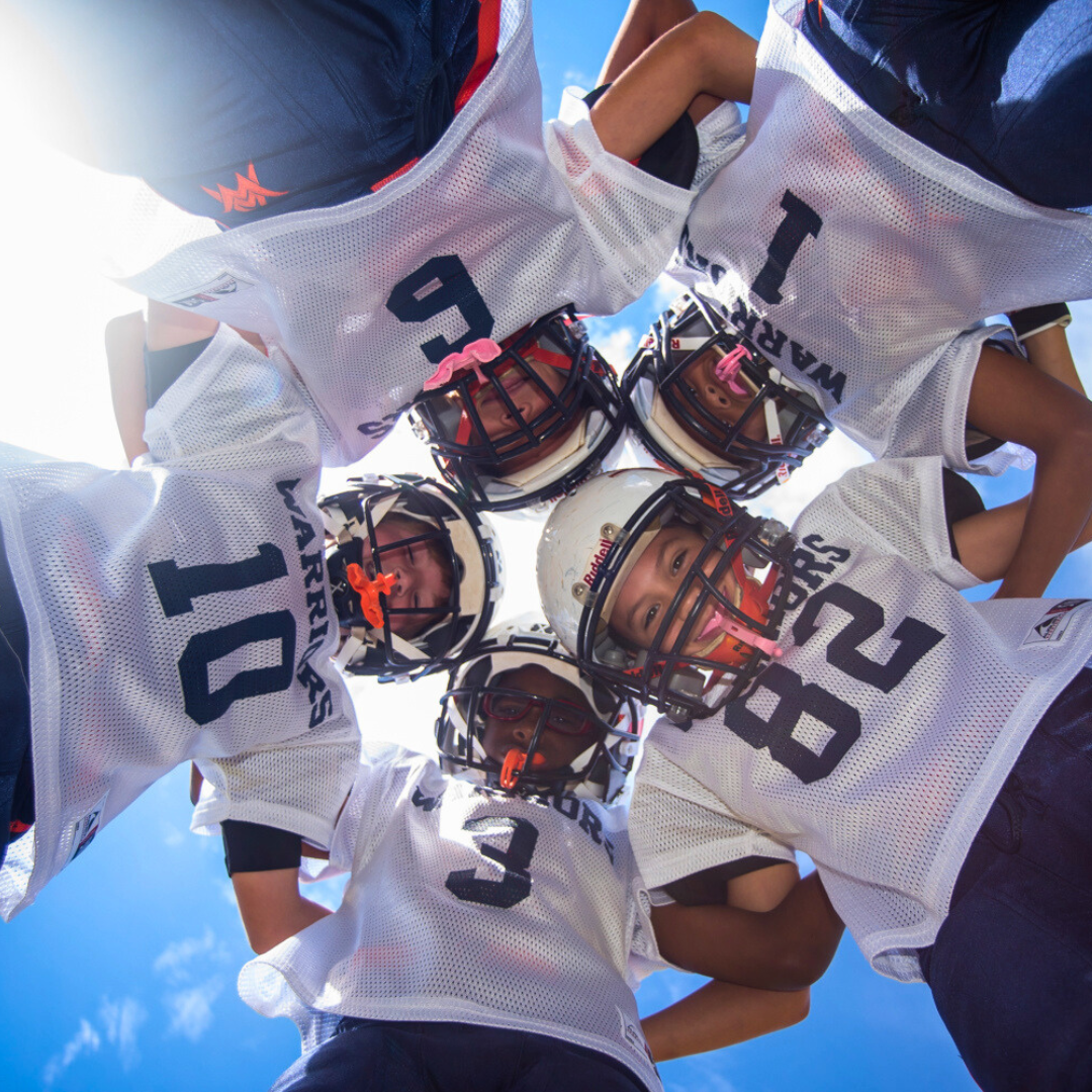 Five youth football players in uniforms and helmets stand in a huddle, looking down at the camera against a bright blue sky, ready to discuss plays and seek additional resources for teamwork.