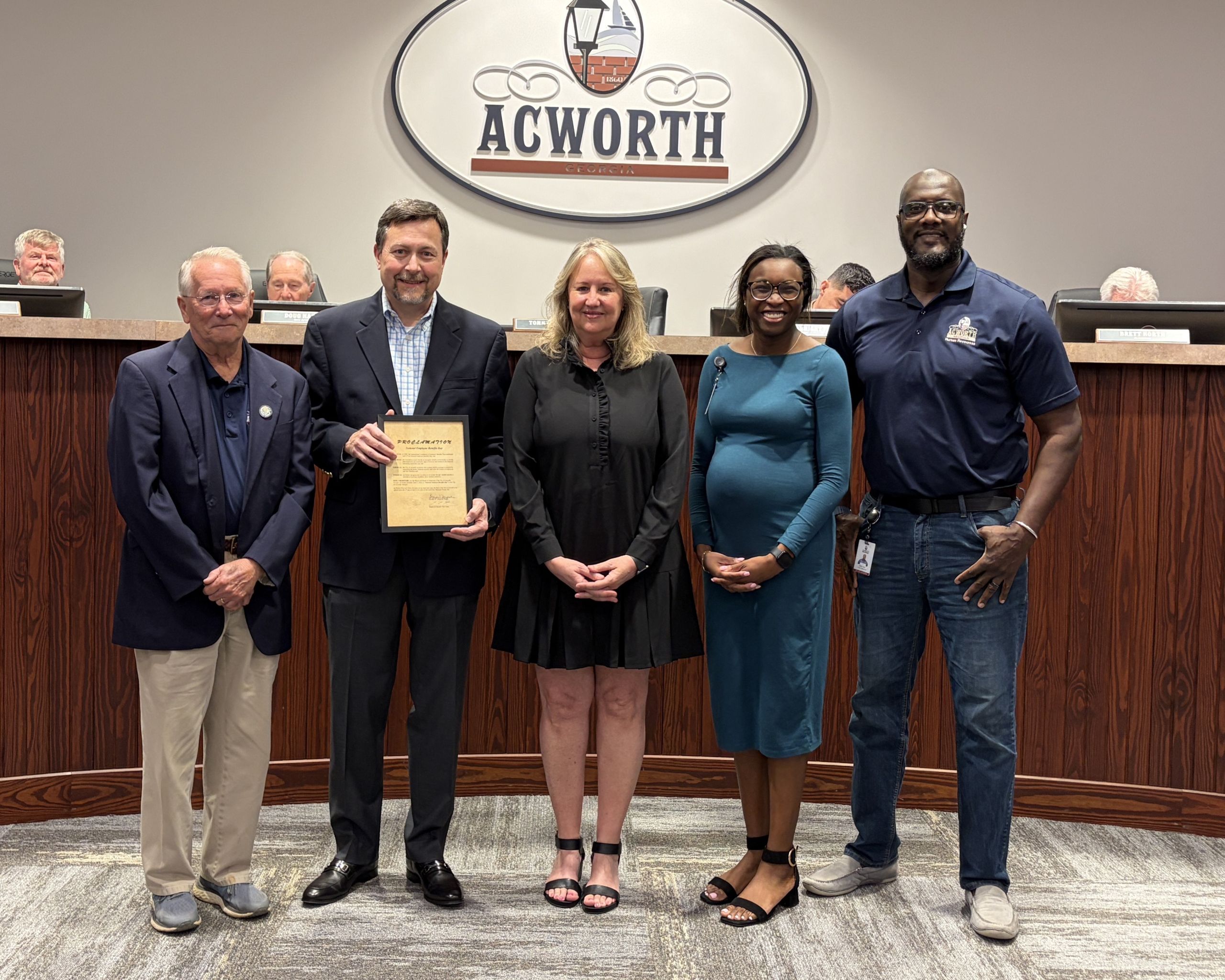 Five people stand indoors in front of an "Acworth" sign; one person holds a plaque, suggesting an award or recognition ceremony at a city council meeting.