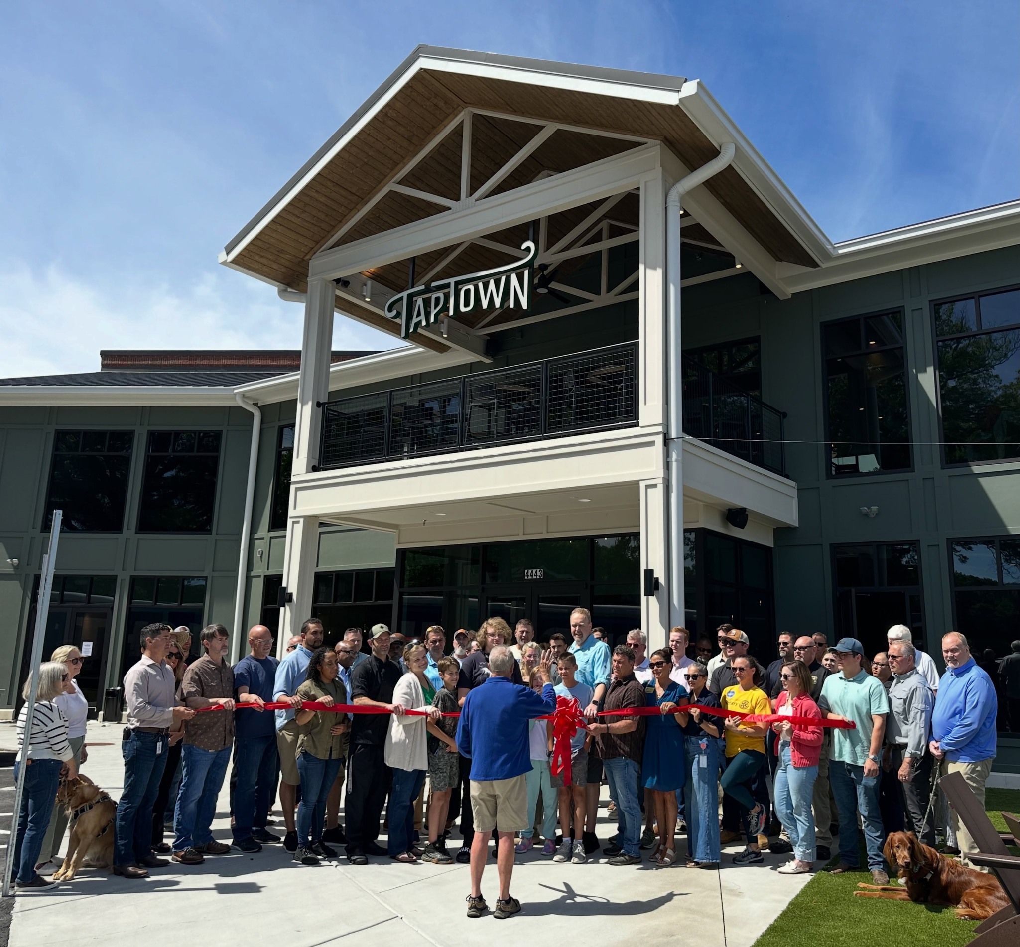 A group of people and several dogs gather outside the TapTown building for a ribbon-cutting celebration, with someone in front holding large scissors.