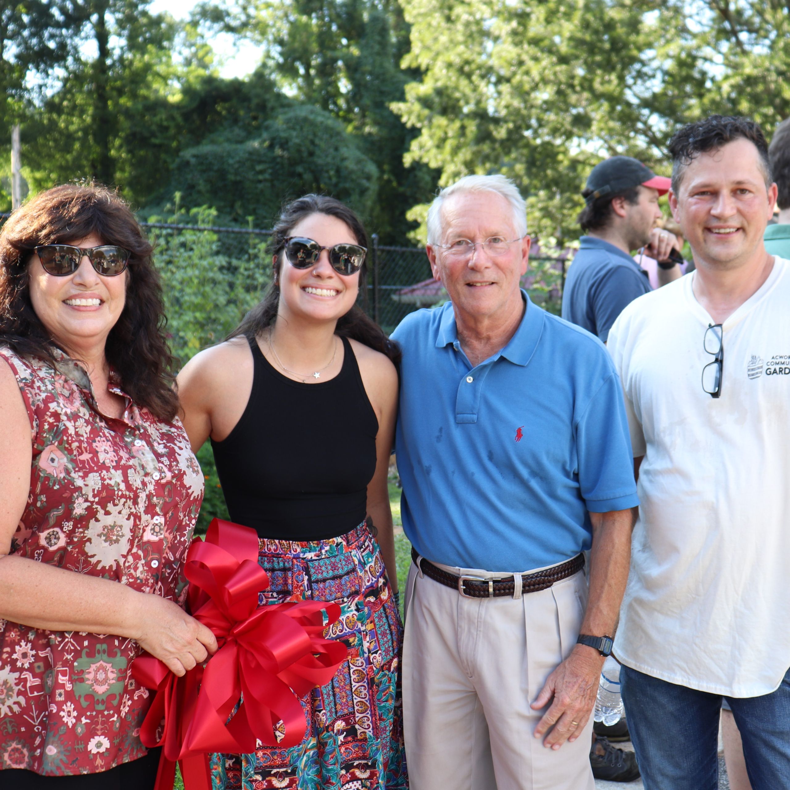 Four people stand outdoors smiling at the camera, one holding a large red ribbon, celebrating the Acworth Community Garden. Trees and fellow community gardening enthusiasts appear in the background.