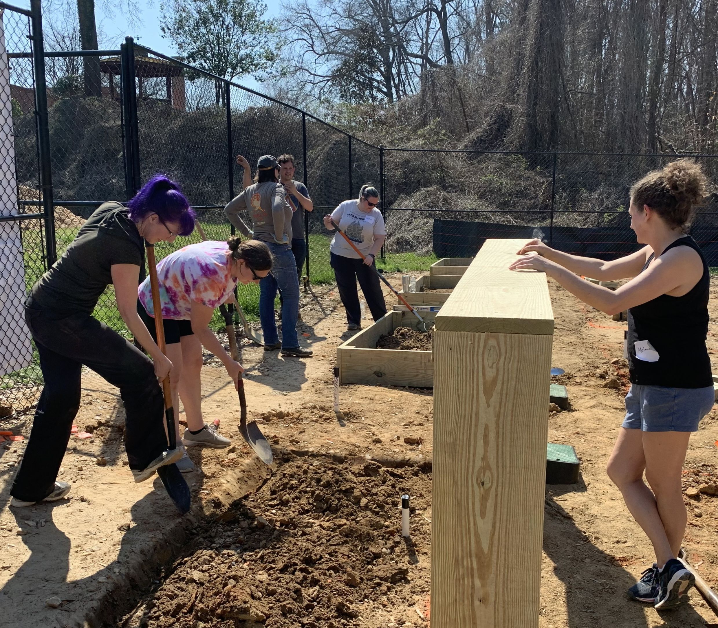Several people work together outdoors at the Acworth Community Garden, digging and moving dirt near a wooden structure, with trees and a fence in the background as part of their community gardening efforts.