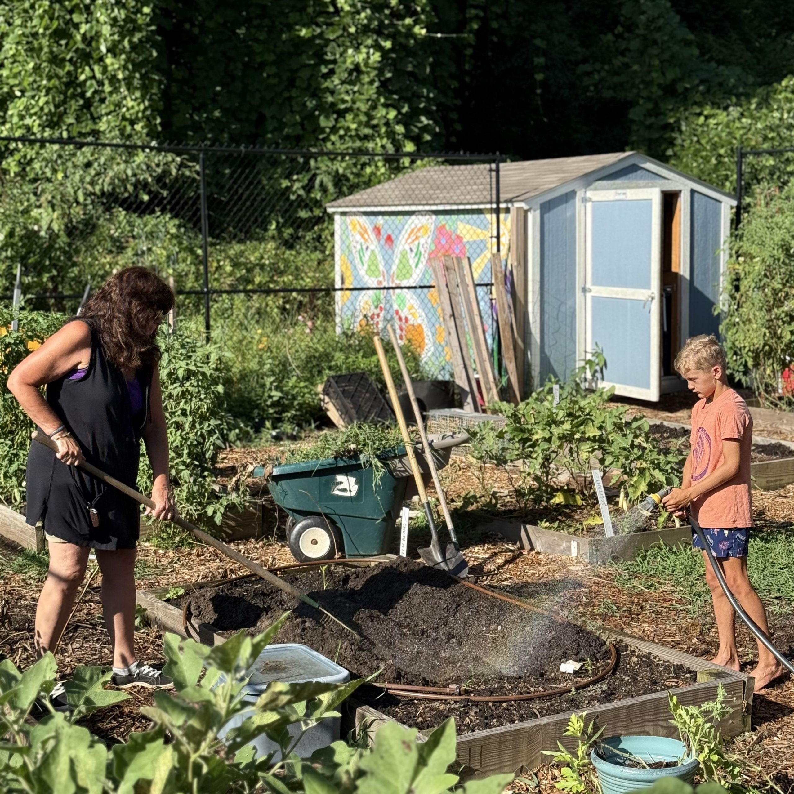 Two people work in the Acworth Community Garden, spreading soil in raised beds to start growing food. A shed with a butterfly mural and garden tools are visible in the background.