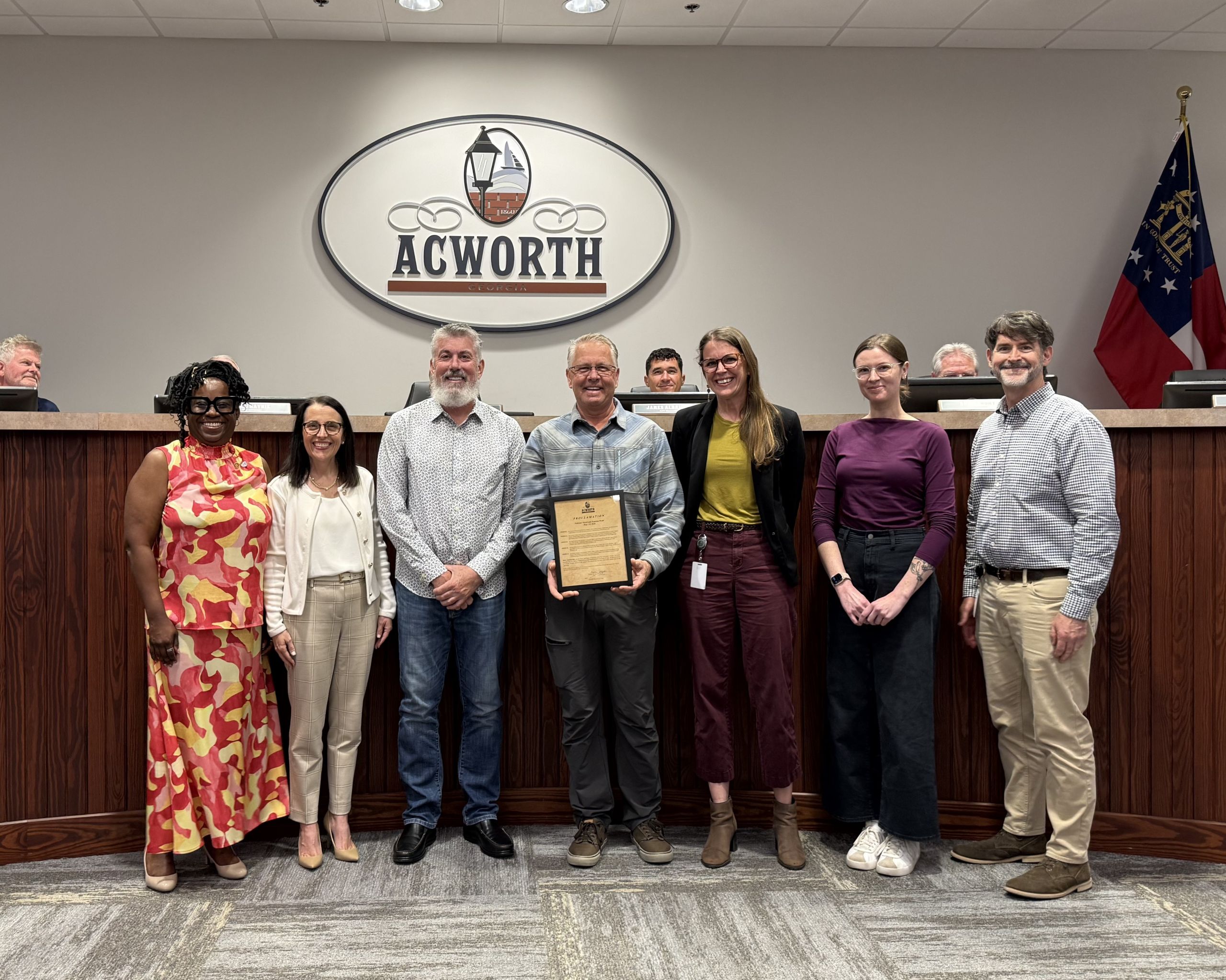 Seven people stand in a row in front of a wooden bench and an "Acworth" sign; the person in the center holds a plaque, likely for recognition or a proclamation during National Travel & Tourism Week. Two flags are visible in the background.