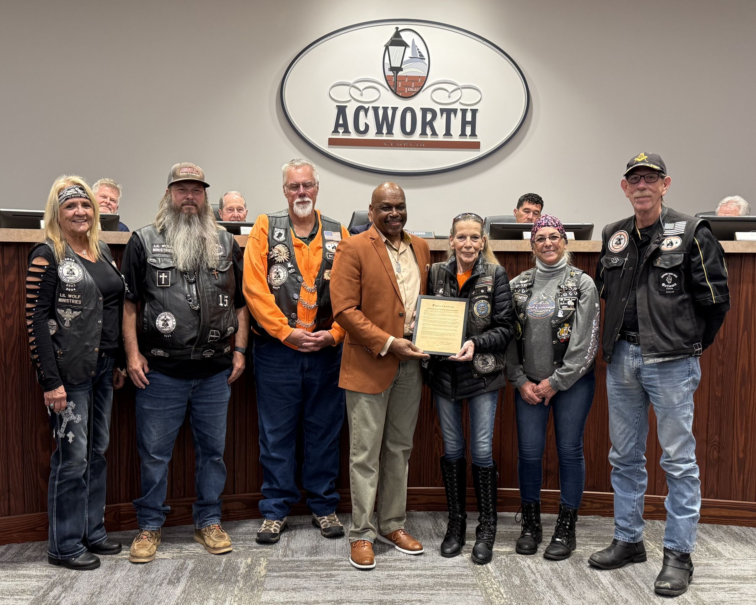 A group of people, some in motorcycle vests, pose with a city official holding a framed certificate in front of an "Acworth" sign at a meeting room, celebrating Motorcycle Safety Month and raising Motorcycle Awareness.