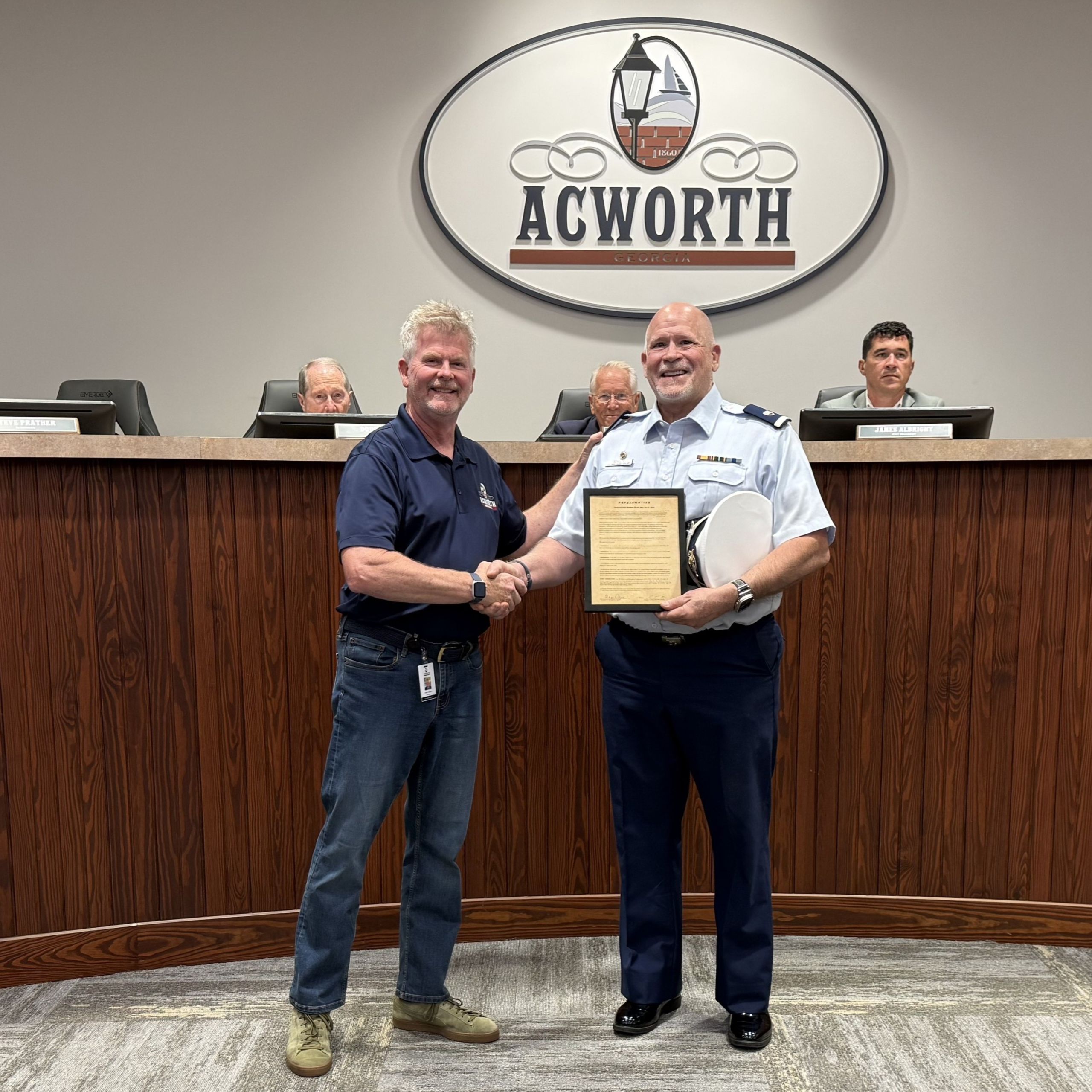 Two men shake hands as one presents an award plaque to the other in front of a city council panel and an "Acworth" sign, celebrating outstanding contributions to boating safety.