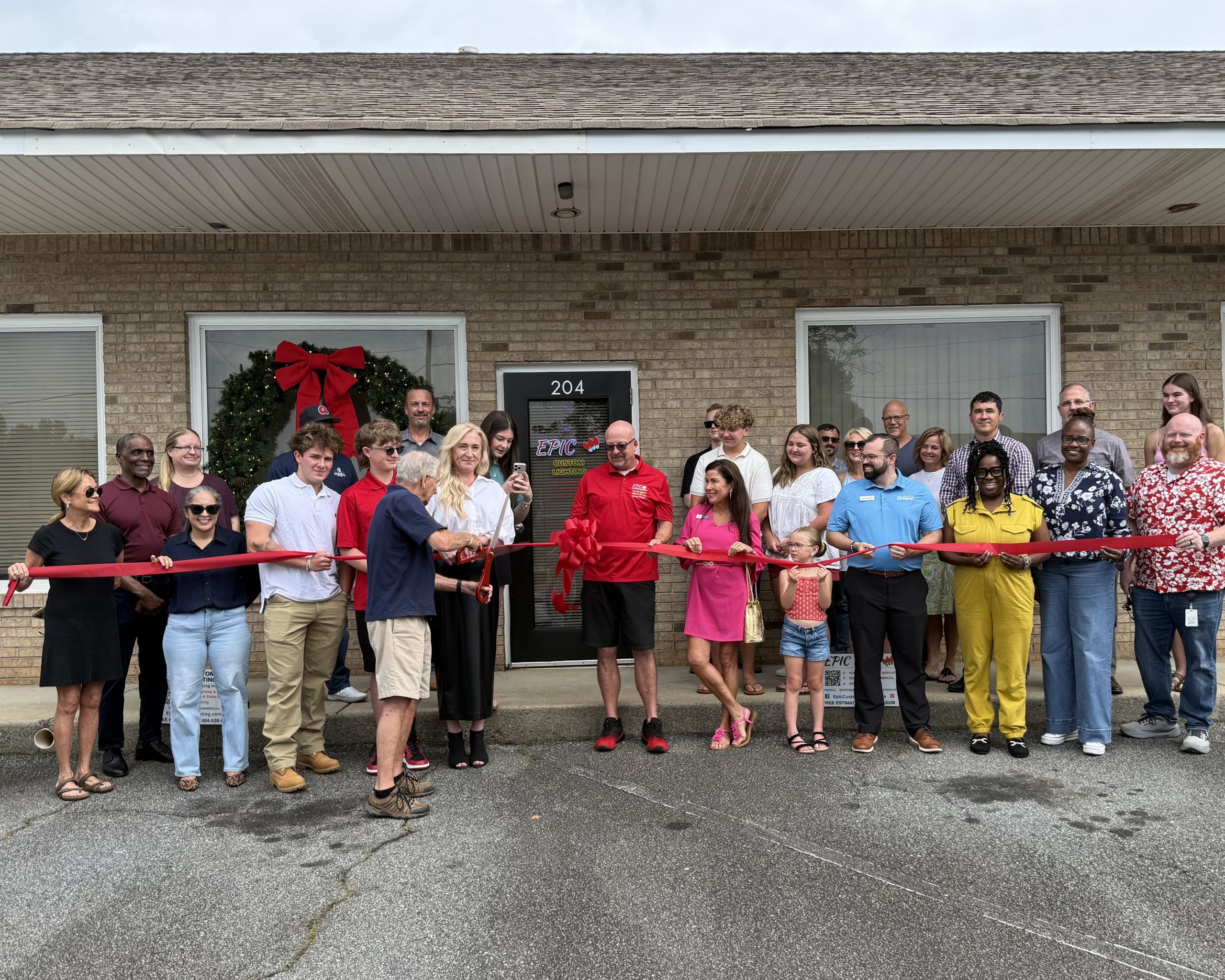 A group of people stand outside a brick building, holding a red ribbon for a Ribbon Cutting ceremony in front of a door labeled "204," illuminated by epic custom lighting.