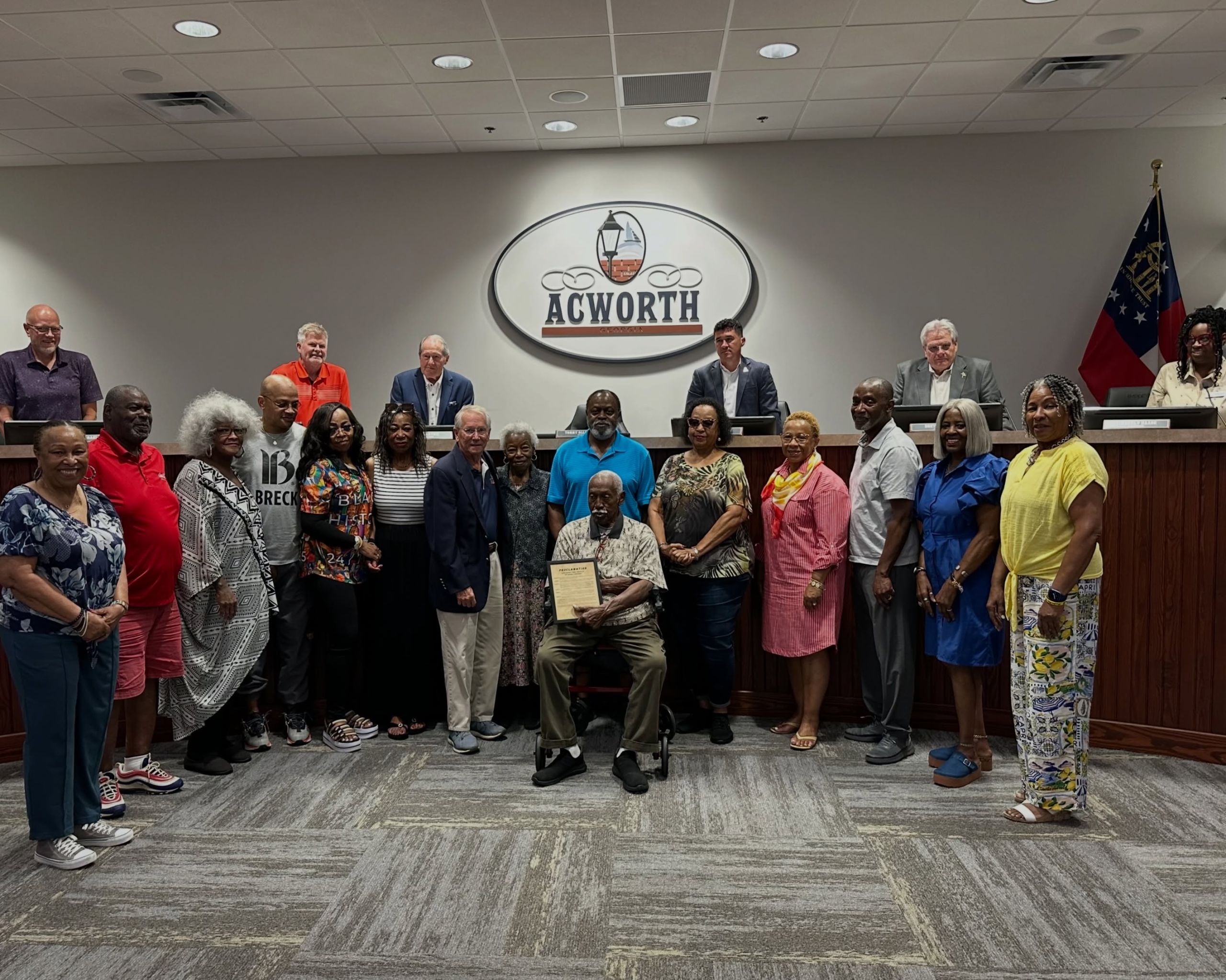 A group of people pose together in a meeting room under an "Acworth" sign, as Claude Johnson is seated holding a plaque and a proclamation celebrating his 90th birthday, with others standing around him.