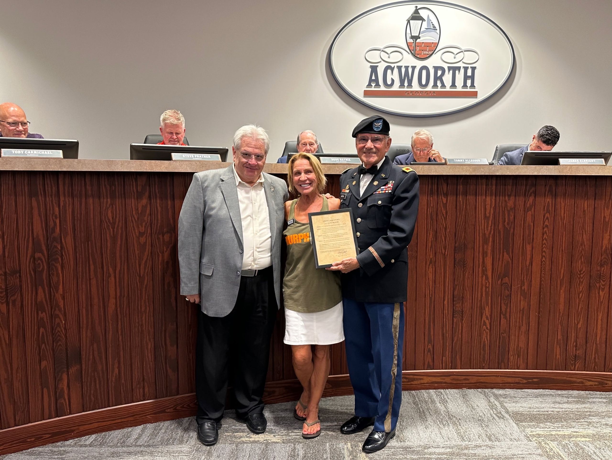 Three people stand in front of a city council bench holding a framed proclamation for US Army Day, with an "ACWORTH" sign on the wall behind them while several officials sit at the bench in recognition of the occasion.