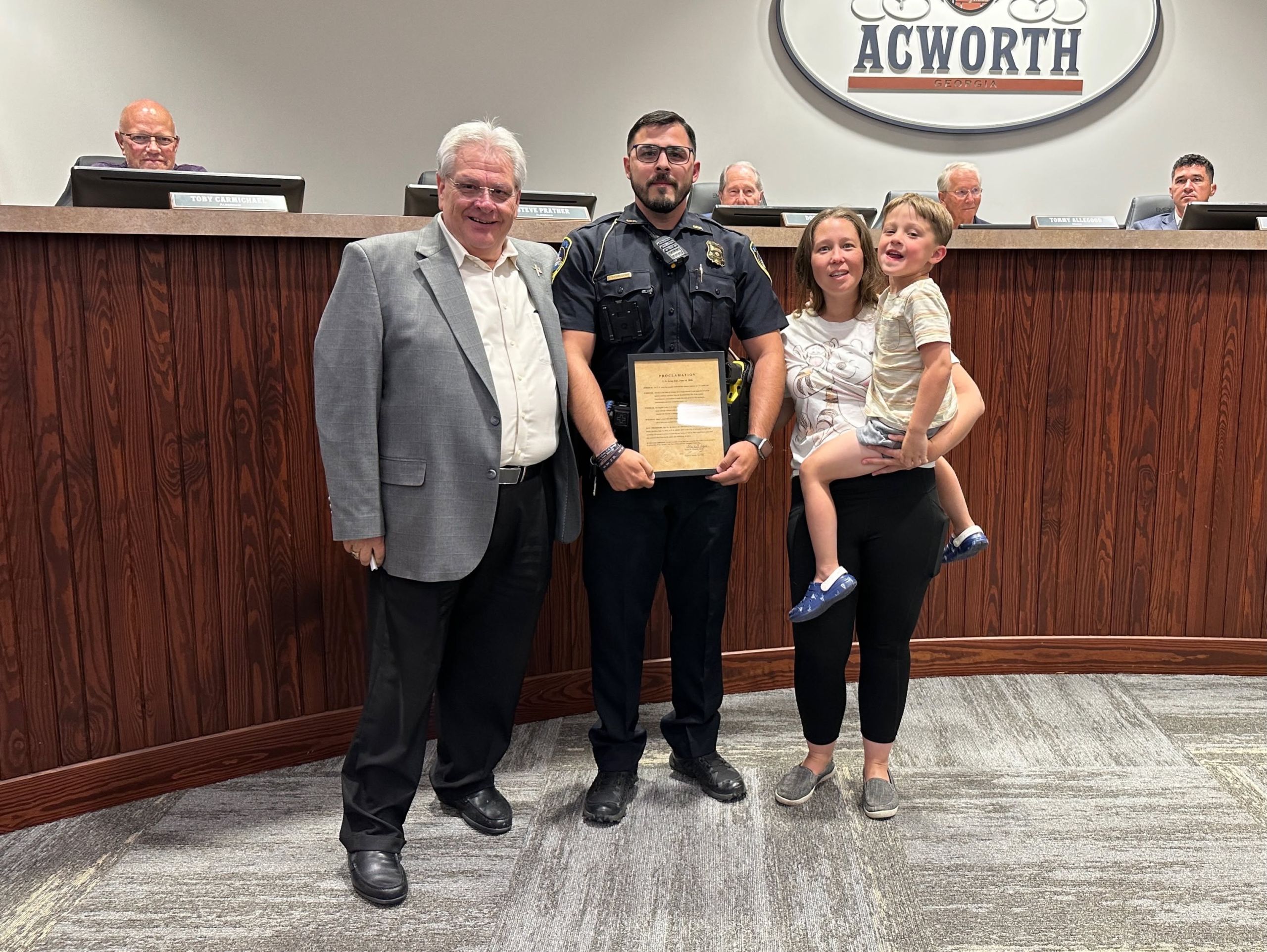 A police officer holds a plaque in recognition while standing with three people in front of a council chamber podium labeled "Acworth," as others sit behind the podium.