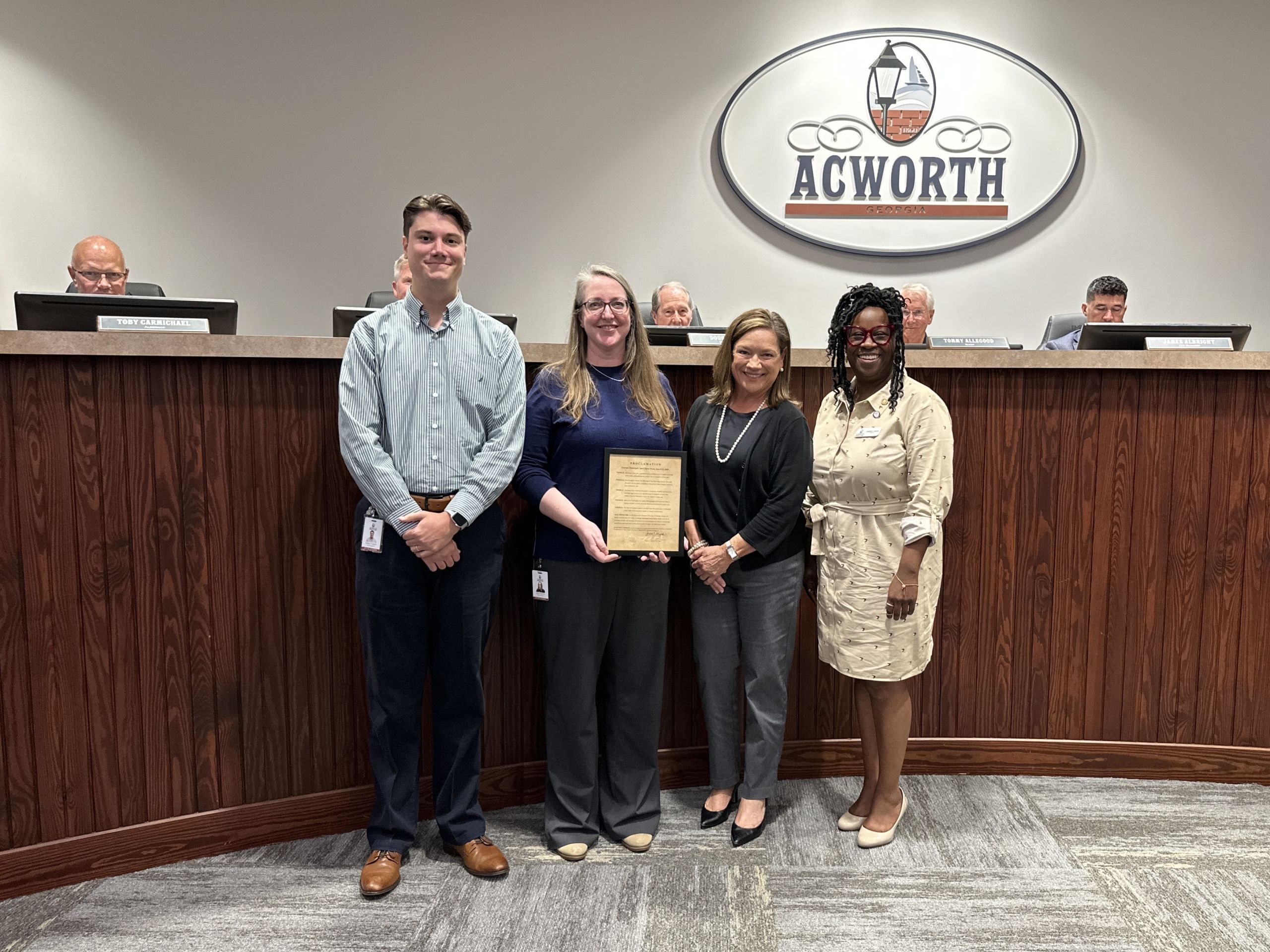 Four people stand in front of a wooden council bench holding a proclamation plaque, with the "Acworth" logo on the wall behind, honoring Municipal Court Clerks Week as three seated individuals look on.