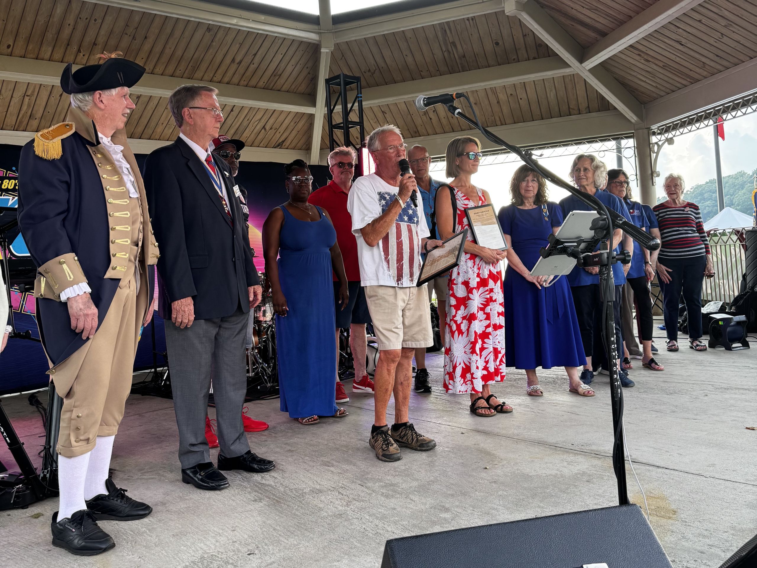 A group of people, some holding papers and microphones, stand on a covered outdoor stage during a public event. One person in historical attire participates in a proclamation honoring the 250th Anniversary of the Declaration of Independence.