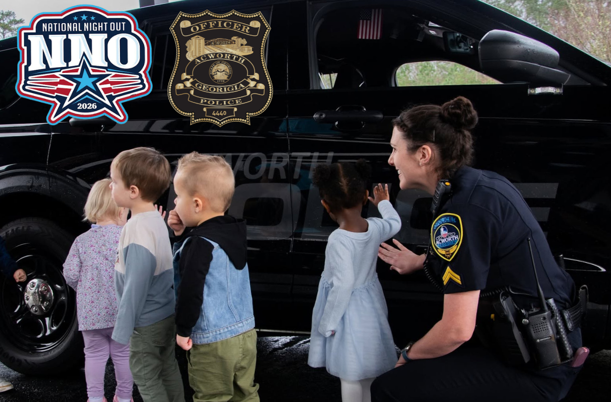 A police officer kneels next to a group of young children, all looking at an Acworth PD Kenworth Police vehicle. National Night Out and police badges are displayed on the car door.
