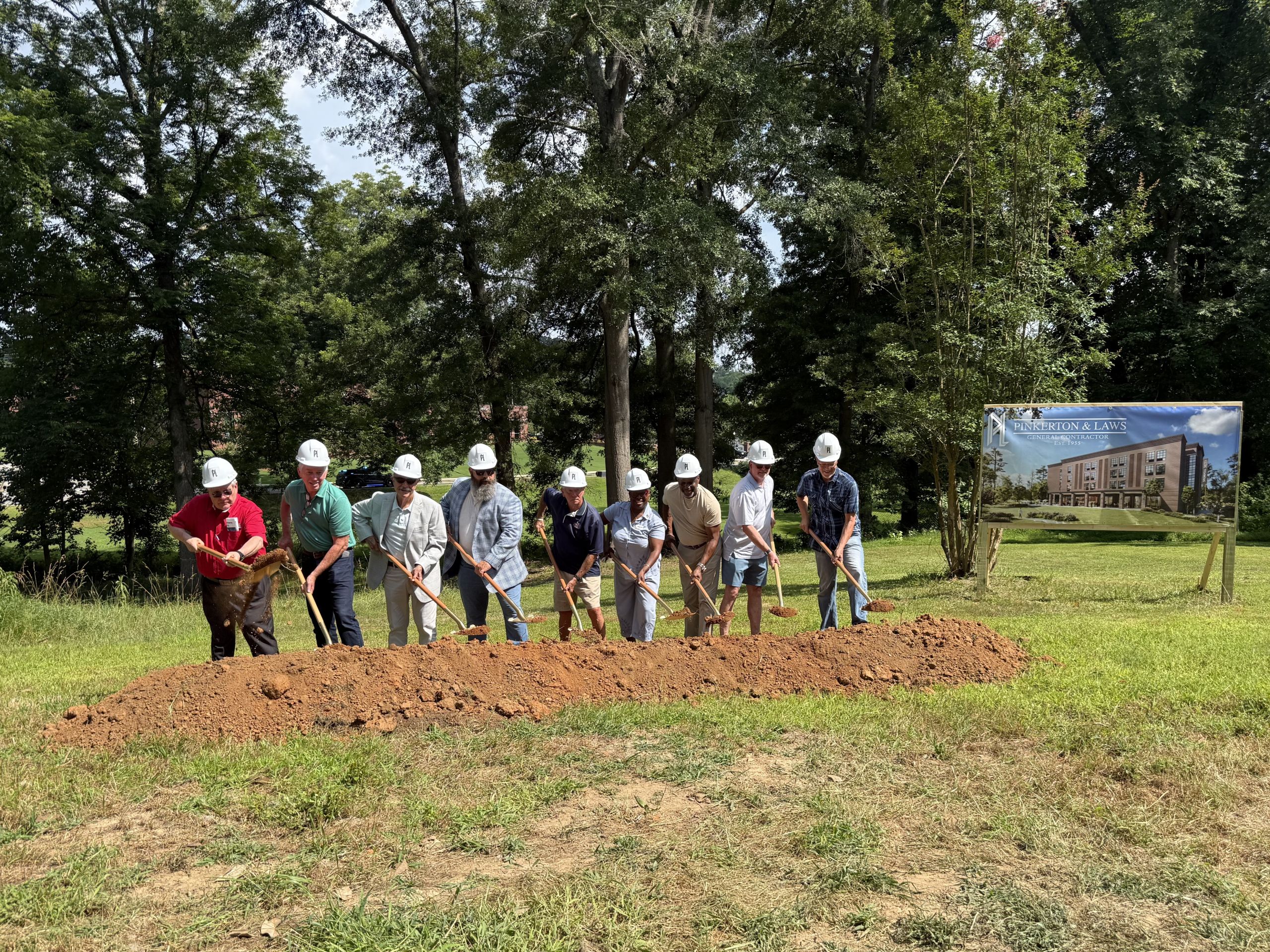 A group of people in hard hats participate in a groundbreaking ceremony for a new downtown hotel in Acworth, standing with shovels in front of a dirt mound and project sign outdoors.