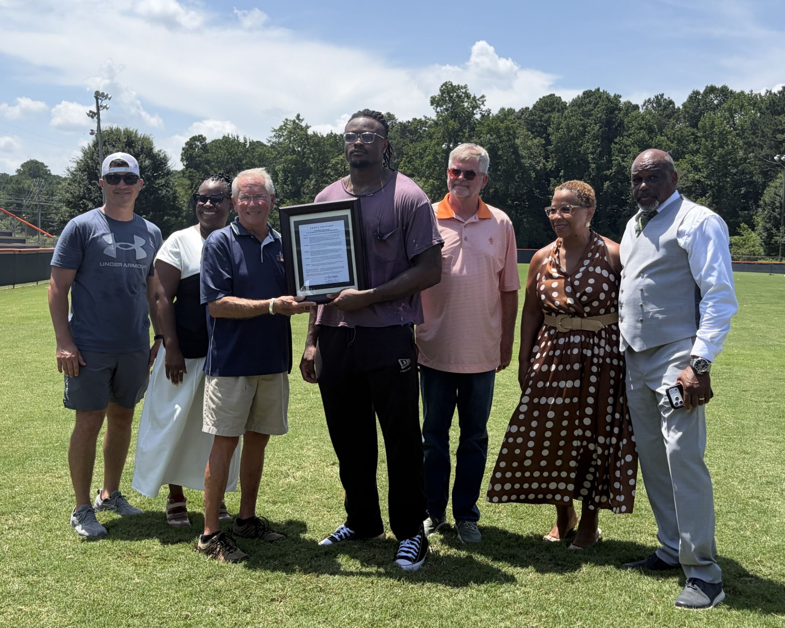 Seven people stand in a row on a field, with one person in the center holding a framed document while shaking hands with another person. Trees and a blue sky are visible in the background.