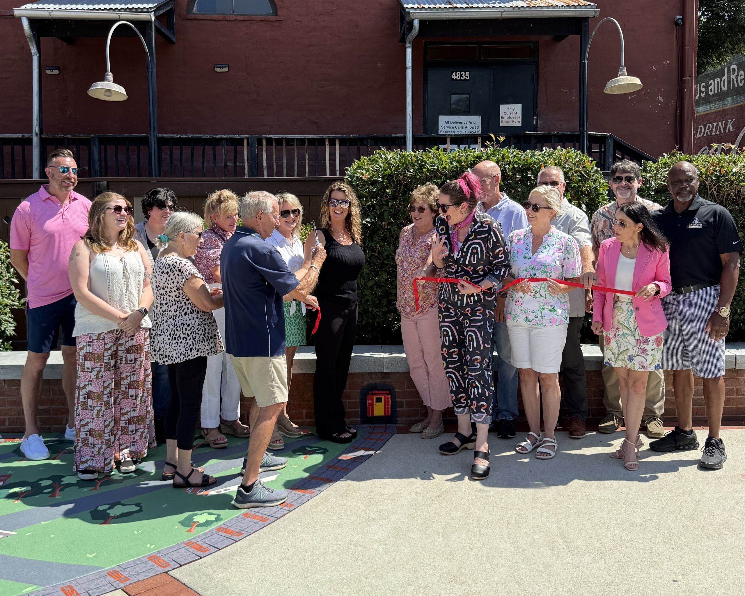 A group of people stands outdoors for a ribbon-cutting ceremony; one person holds oversized scissors, while others watch and smile.