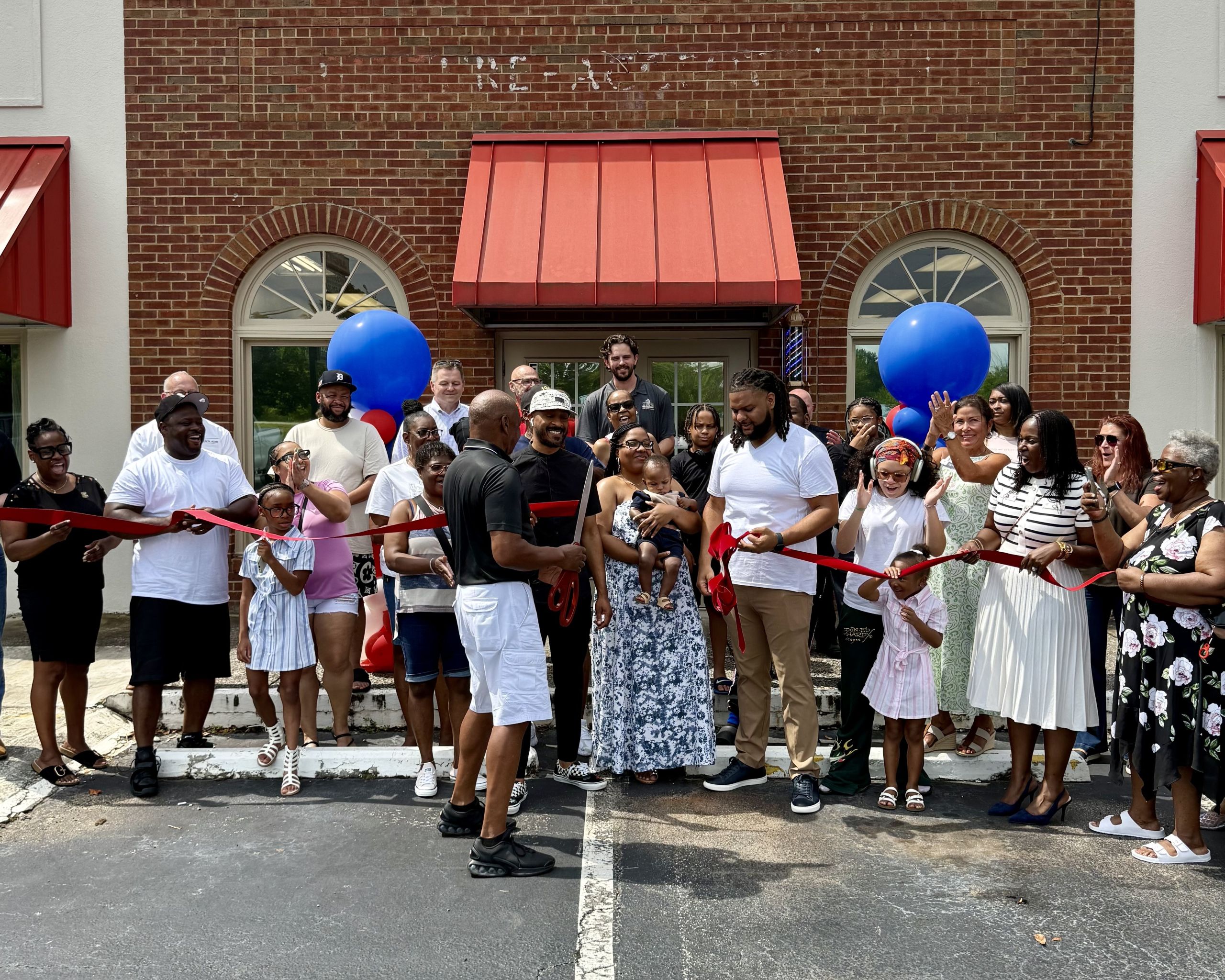 A group of people stand outside a brick building with red awnings, holding a large red ribbon for the Gifted Barber Shop ribbon-cutting ceremony; two people hold oversized scissors in the center.