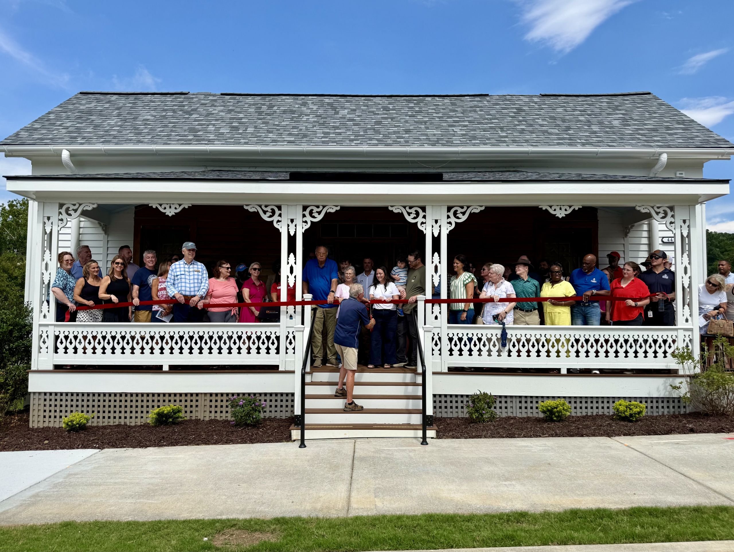 A group of people stands on the porch of the McCray Farmhouse during a ribbon-cutting event, celebrating its recent restoration on a sunny day.