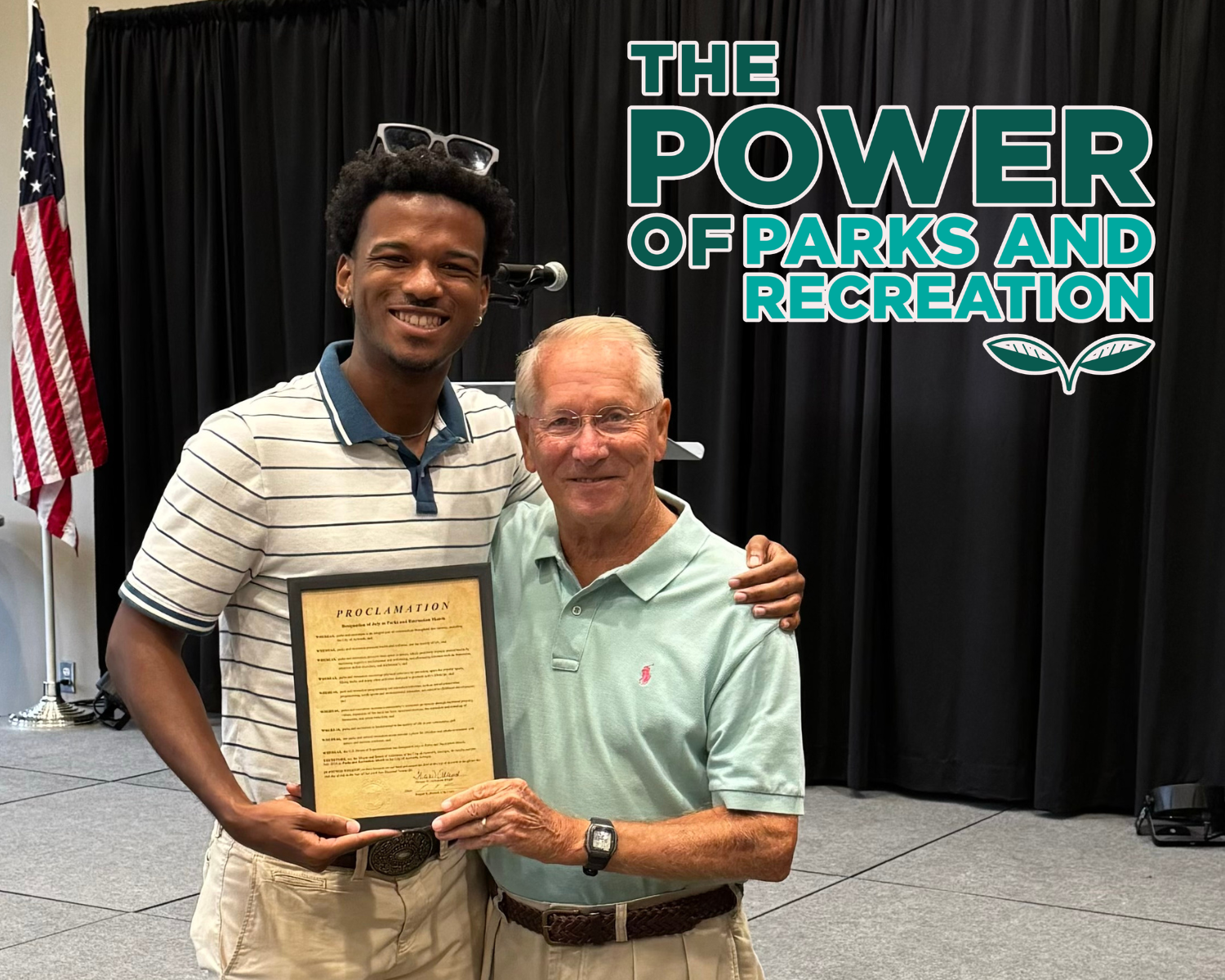 Two men stand together smiling, holding a framed proclamation celebrating Parks and Recreation Month. Behind them is a black curtain, U.S. flag, and a sign that reads “The Power of Parks and Recreation.”.