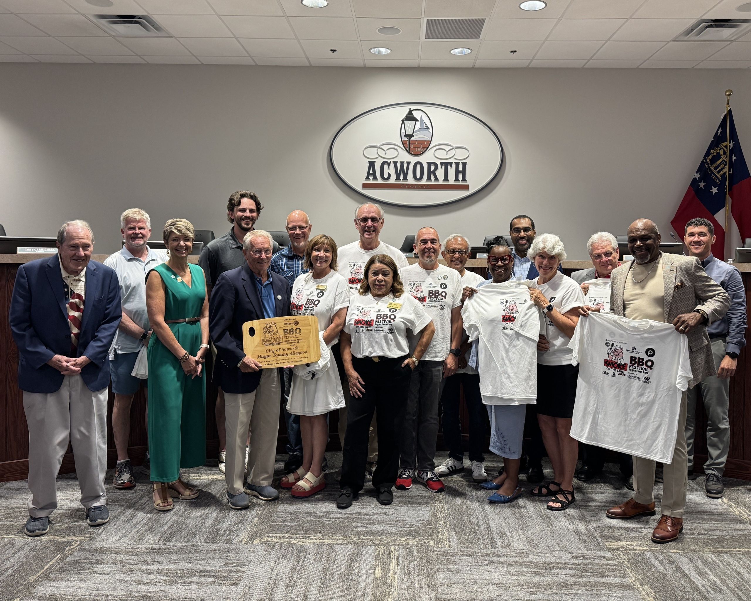 A group of people pose together in a room under an Acworth sign, proudly holding BBQ event t-shirts and a commemorative plaque from the 2026 North Cobb Rotary Smoke on the Lake BBQ Festival.