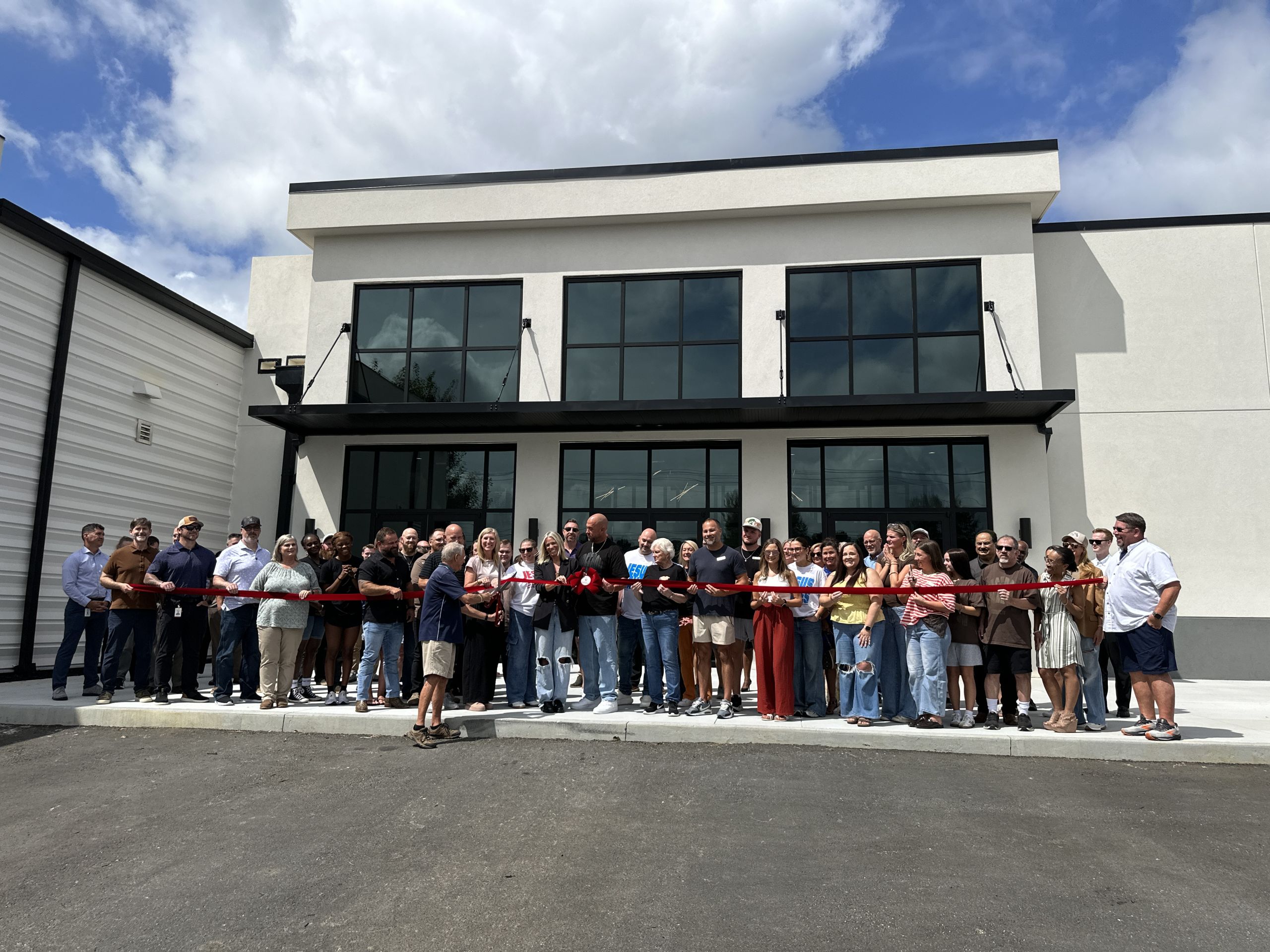 A large group of people stands outside the modern Freedom Church building, holding a red ribbon for the New Auditorium ribbon-cutting ceremony under a partly cloudy sky.
