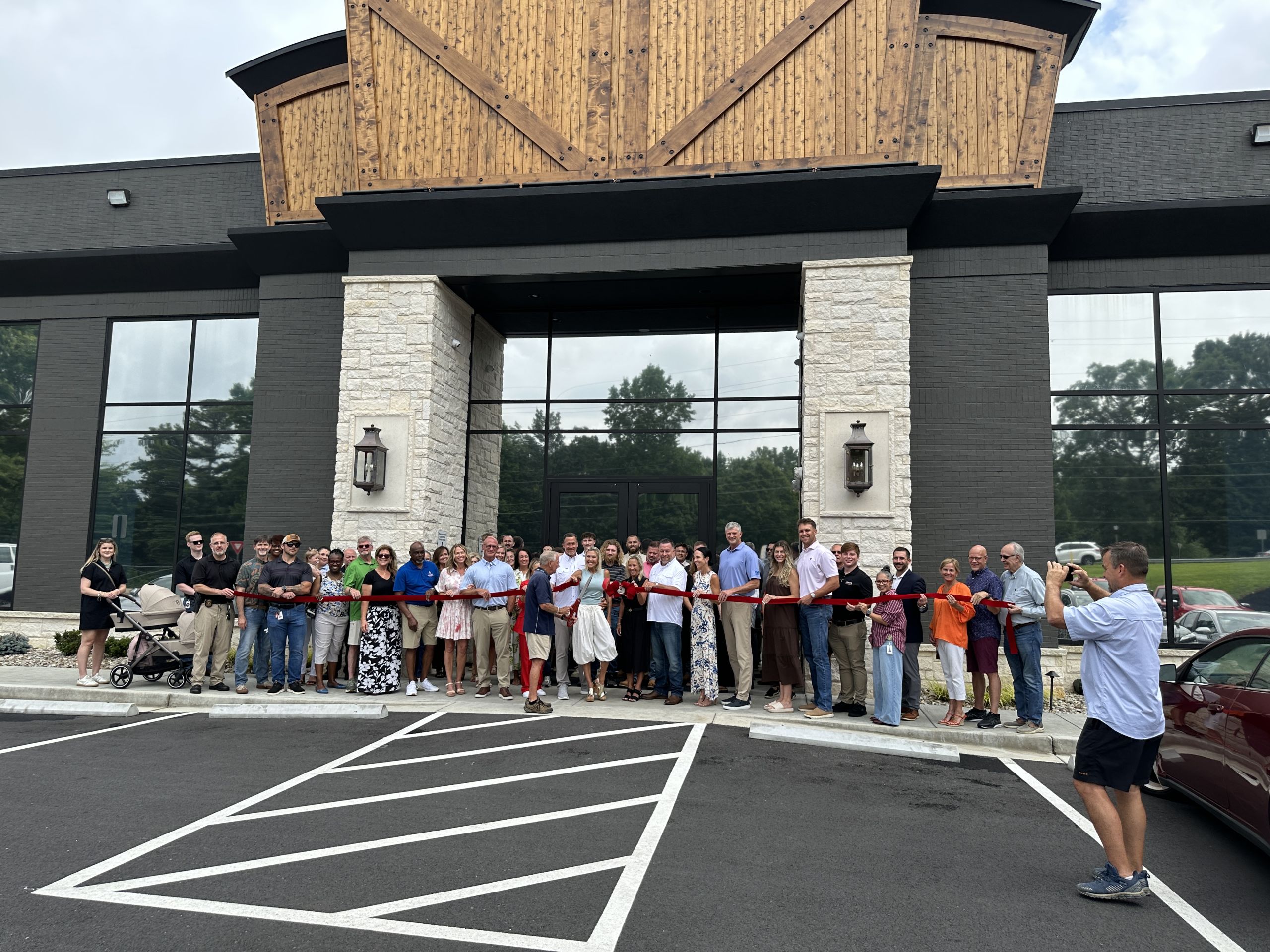 A group of people stand in front of a building for a ribbon cutting ceremony celebrating the new headquarters, while one person takes their photo in the parking lot.