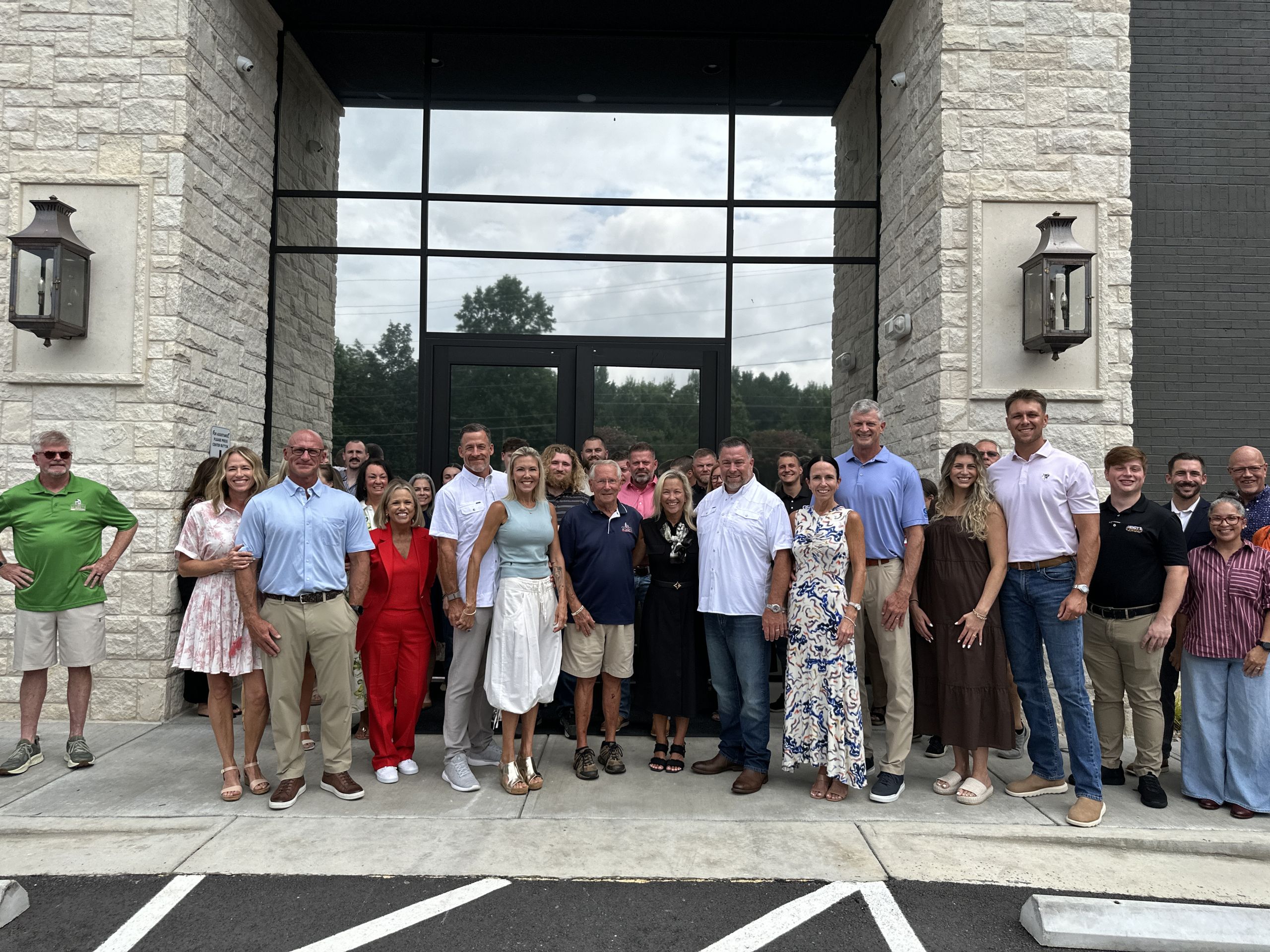 A group of about 25 adults pose together outside a building with stone walls, large glass doors, and lantern-style lights on either side, celebrating the ribbon cutting of the new headquarters alongside fellow local businesses.
